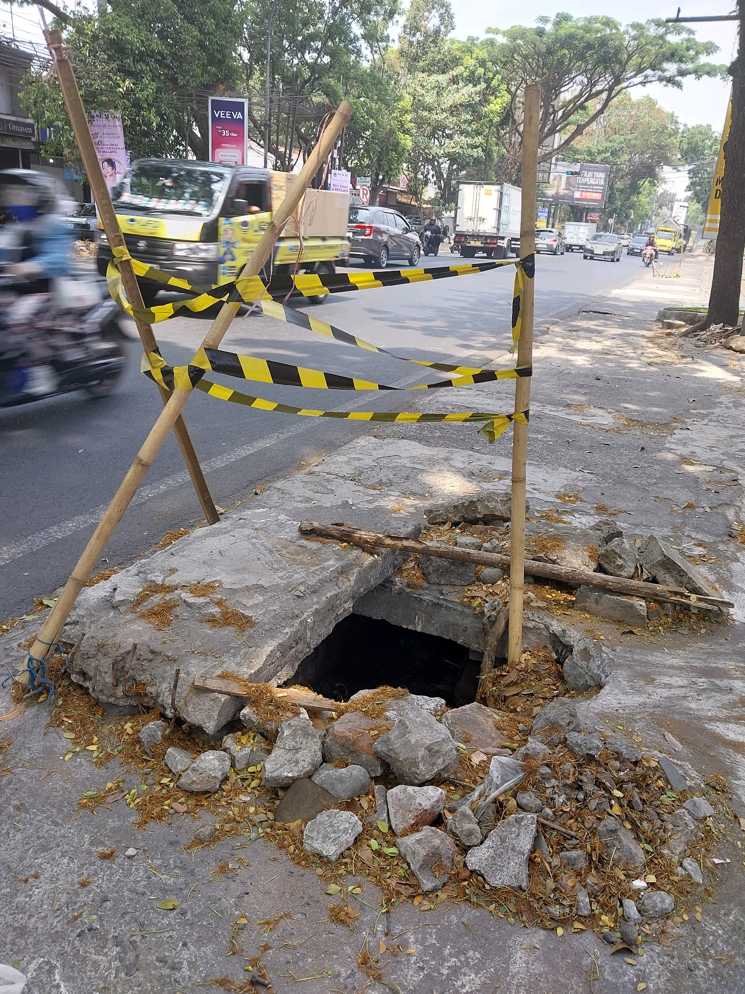 This is an eye-level, medium shot of a hole in a concrete sidewalk, cordoned off with yellow and black caution tape supported by bamboo poles. The hole appears to be a manhole or drain, and it is surrounded by loose concrete debris and fallen leaves. In the background, a busy street in the city of Malang, Indonesia, is visible. Cars, trucks, and a motorcycle are driving on the road, with trees and buildings lining the street. The overall scene depicts a construction or maintenance hazard on a public thoroughfare.