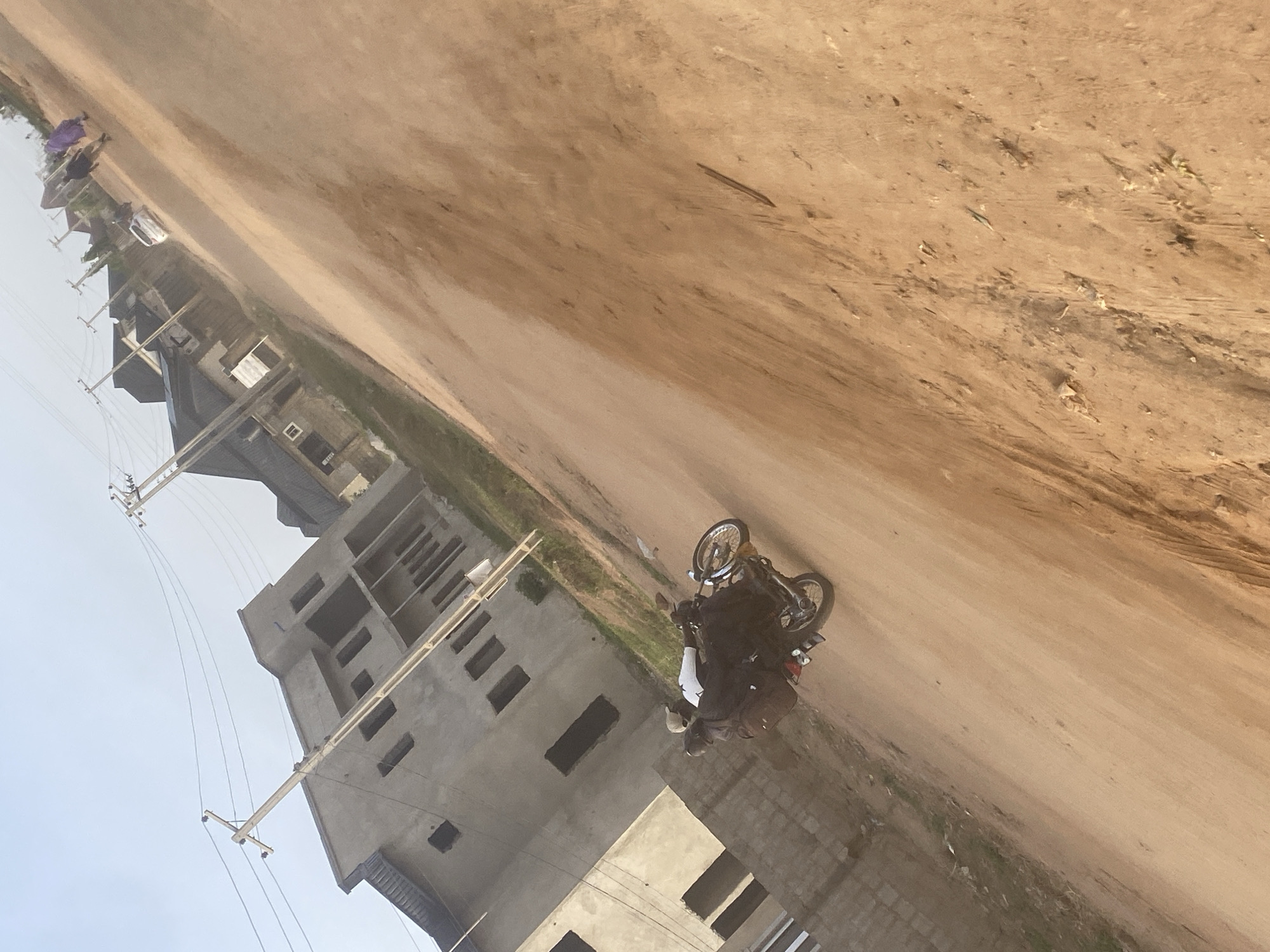 The image shows a street scene in Lugbe, Nigeria. A dirt road runs diagonally across the frame. A motorcycle with a rider is situated on the road. To the left are buildings, some under construction, with electrical poles and lines. People are present further down the road towards the left. The sky is visible in the upper left corner.