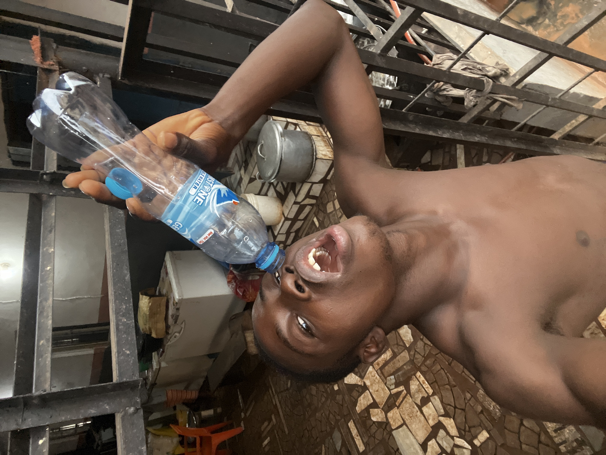 In Nkpor, Nigeria, a young shirtless Black male is shown from a low-angle perspective, his mouth wide open as he attempts to drink from a nearly empty plastic water bottle held upside down. The bottle's opening is positioned directly over his nose and mouth. He is outdoors, with a patterned tiled floor visible beneath him. In the background, there is a metal railing, a white freezer, an orange plastic chair, and several pots. The lighting suggests it is daytime.