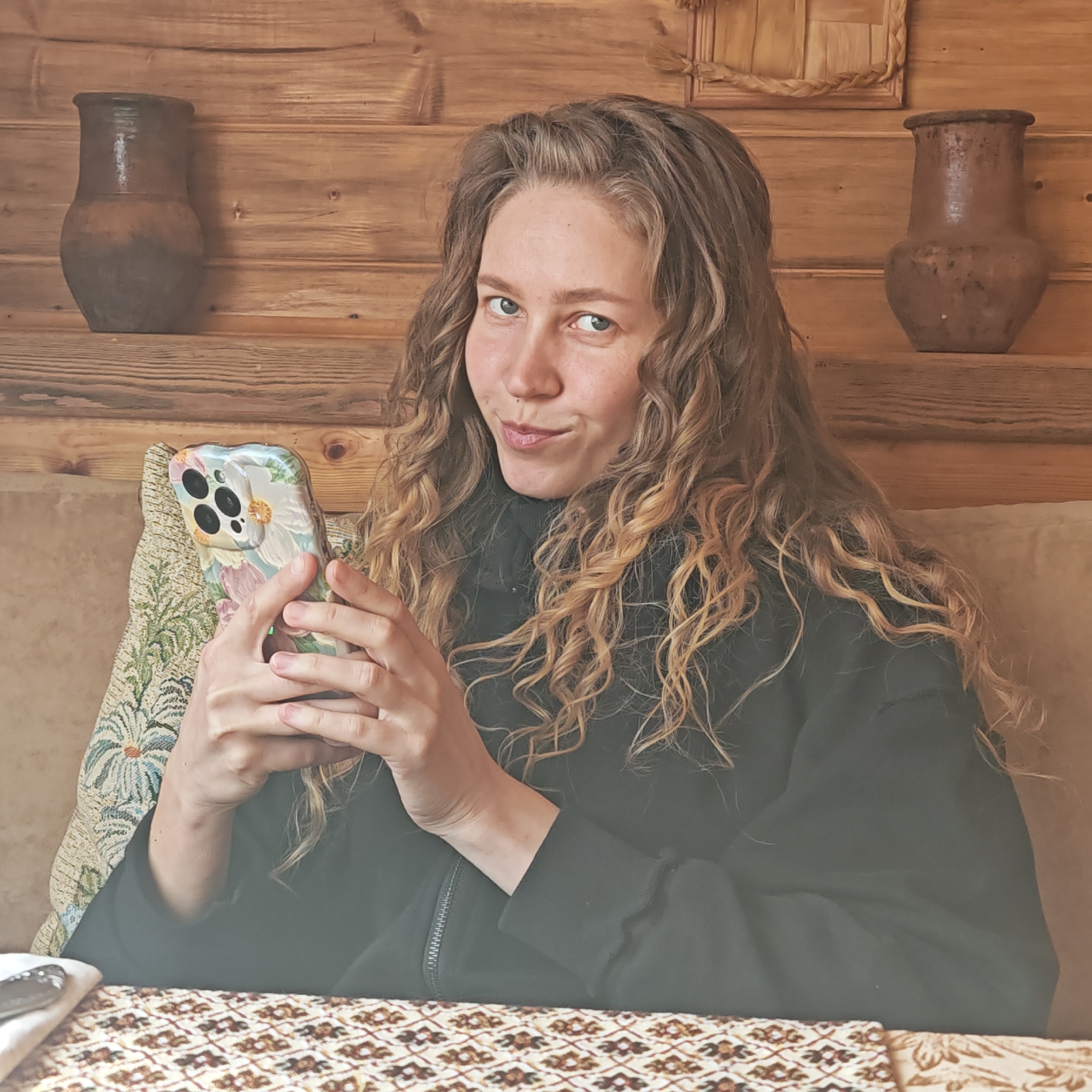 A young woman with long, wavy hair is seated at a table in what appears to be a rustic restaurant or dining area. The location is Rostov, Russia. She is holding a smartphone with a floral design case. The background features wooden plank walls with two earthenware pots displayed on a shelf. The table is covered with a patterned cloth, and a pillow with floral embroidery is visible behind her. She is wearing a dark-colored zip-up sweater.