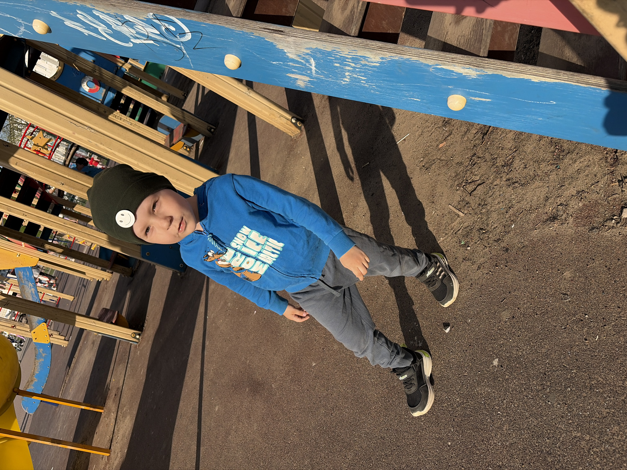 A boy stands on what appears to be a playground, near a climbing structure made of wood and painted blue, yellow, and pink. He is wearing a dark green beanie with a white circular patch, a blue zip-up hooded sweatshirt, grey pants, and black and white sneakers. The playground surface is brown, and there are shadows cast from the playground equipment and the boy himself. The playground is likely located in the city of Chaikovsky, Russia, given that the prompt requests the city.