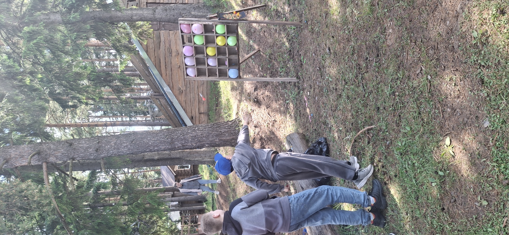 A wide shot of a forest scene in Прикамский, Russia shows a man in grey clothing and a blue cap in the process of throwing a dart toward a wooden balloon dart game with pink, green, yellow and blue balloons. Another person is laying on the ground behind him, and another person is standing in the background to the left. A wooden structure is to the left of the dart game, and there are scattered trees. The ground is covered with grass and pine needles.