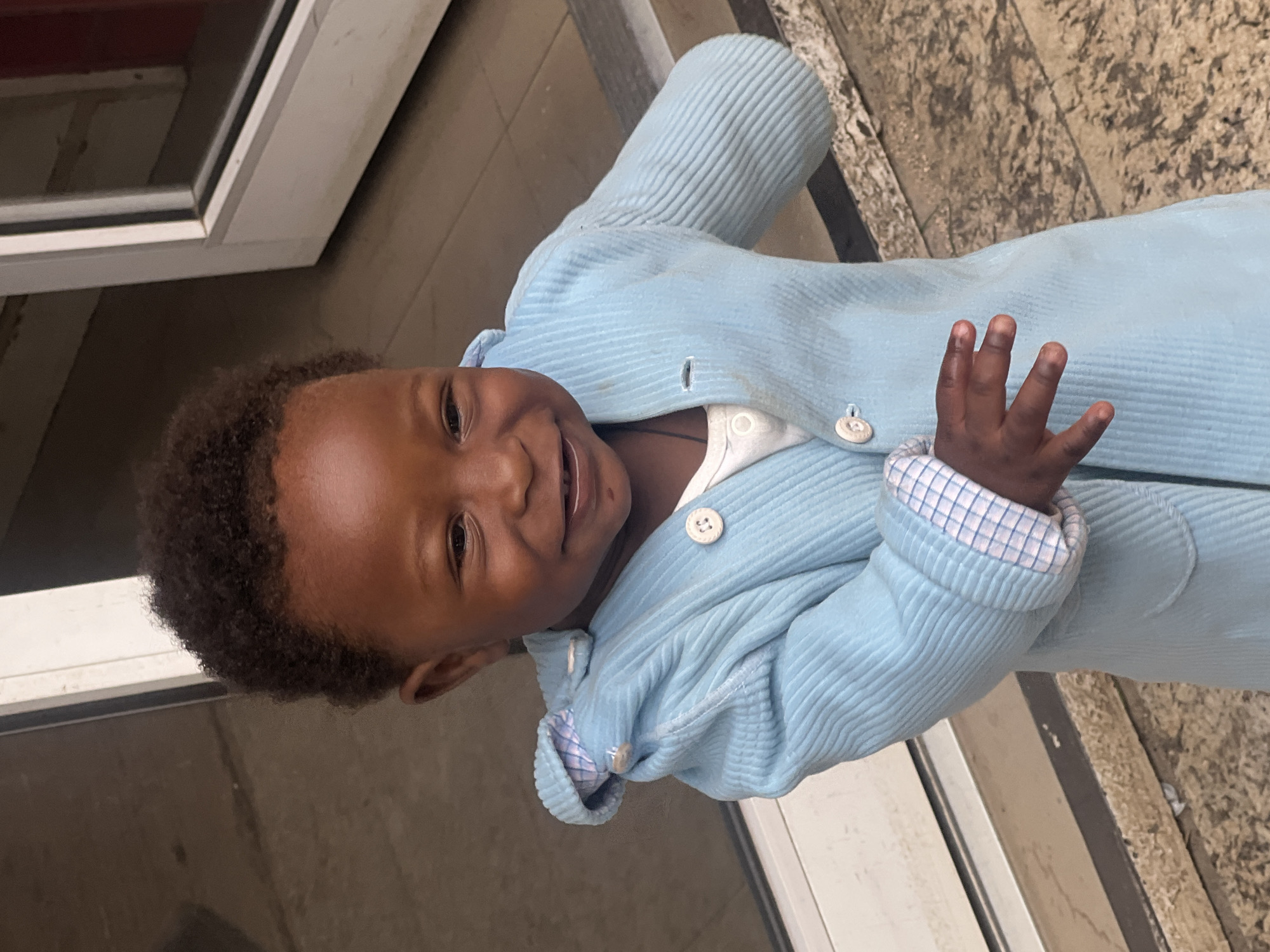 A close-up photograph shows a young child with dark, curly hair, light brown skin, and dark eyes. The child is smiling and looking up at the camera, with their right hand raised, fingers spread slightly. The child is wearing a light blue corduroy jacket over a white undershirt. The jacket has white buttons and a blue and white checkered lining visible at the cuffs and hood. The background consists of a light-colored, possibly granite, surface and a white door frame. There is no indication of the city of Gura Top, Nigeria.