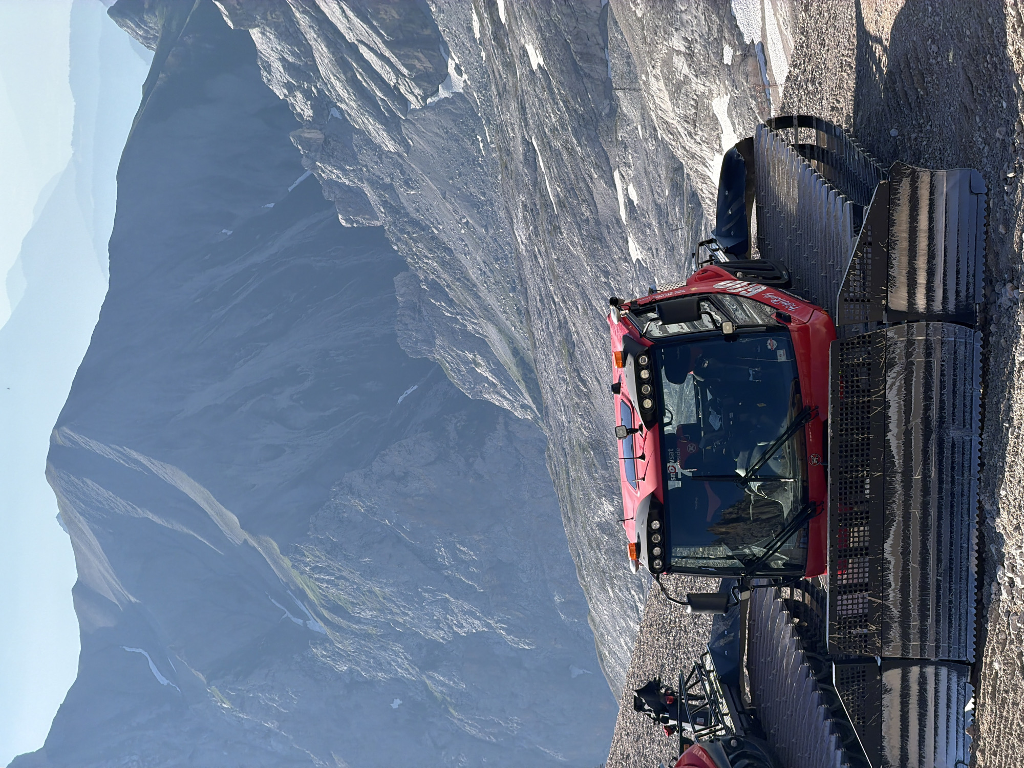 This media file shows a bright red snow groomer parked on a rocky, mountainous terrain. The snow groomer has large, black tracked wheels and a prominent cabin with windshield wipers. Inside the cabin, at least one person is visible, appearing to be the driver. The background reveals a vast, dramatic mountain landscape with steep, rocky slopes and patches of snow. The lighting suggests it is daytime, with the sun casting shadows on the ground and highlighting the texture of the mountains. The sky transitions from a bright blue near the horizon to a hazy, lighter blue at the top, indicating a clear or partly cloudy day. The steepness and rocky nature of the terrain suggest a high-altitude location, possibly in the Alps. Text is visible on the snow groomer, including "Snowcat" and "Garmisch-Partenkirchen", confirming the location. The scene appears serene and adventurous, with the powerful machine poised against the rugged natural environment.