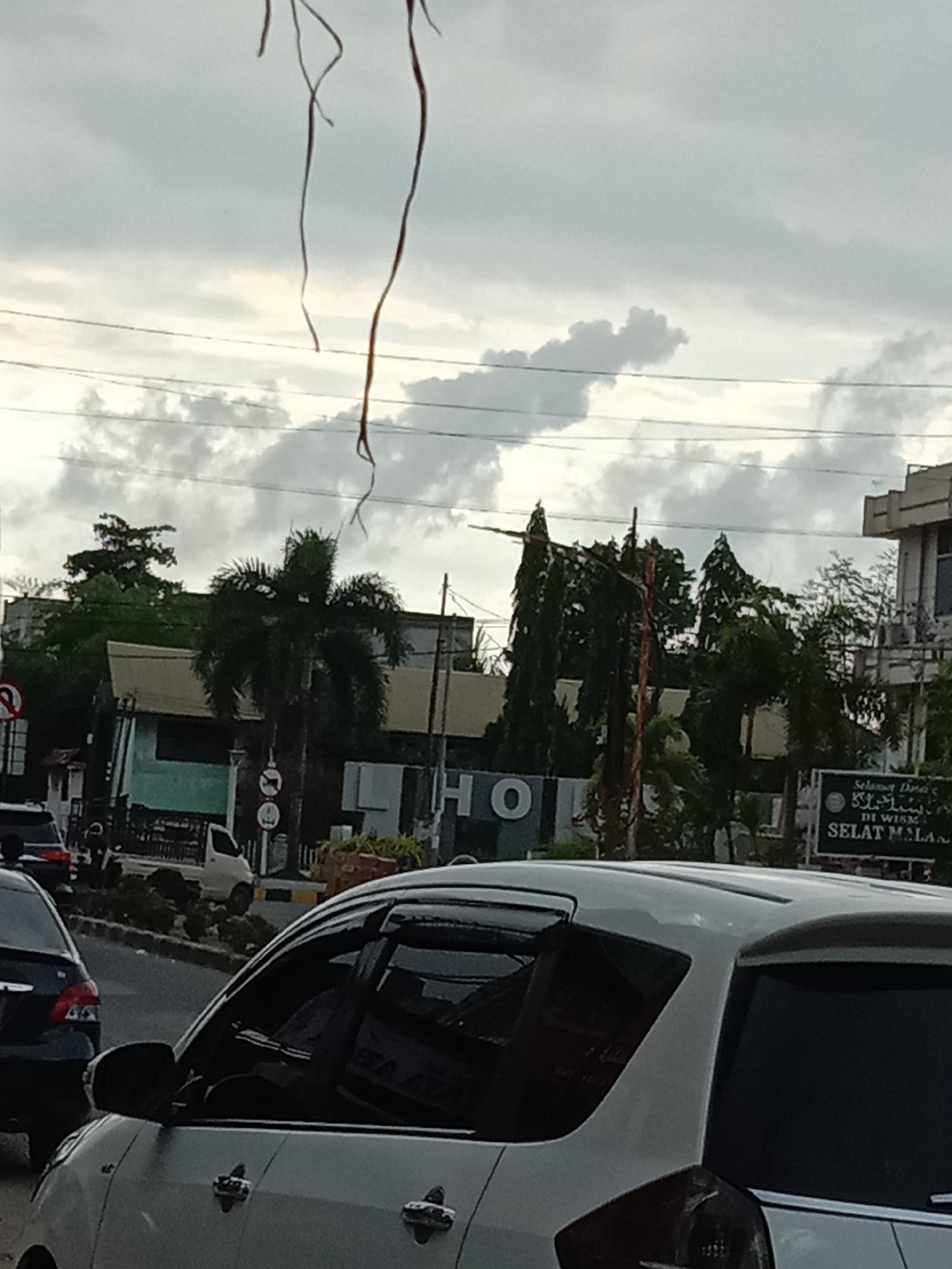The image captures a street scene in Lhokseumawe, Indonesia under an overcast sky. In the foreground, a white car is partially visible, occupying the lower portion of the frame. To the left, the rear of a dark-colored vehicle is also present.
Behind the cars, there is a road with other vehicles and pedestrian walkways with greenery. Further back, buildings are visible, including one with the letters "HO" displayed. The street signage reads "Selamat Datang DI WISMA SELAT MALAN". Trees, including palm trees, are interspersed among the buildings.
In the upper portion of the image, utility wires stretch across the sky, and stringy vines are seen hanging in the foreground obscuring part of the scene. The clouds are dense and gray.