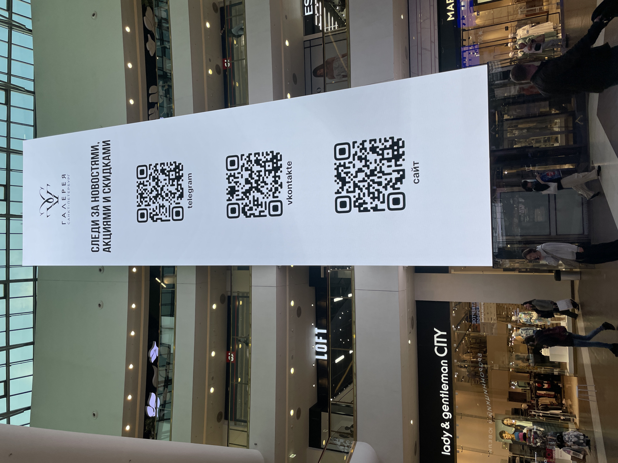 Here is a detailed description of the image:
**General Scene:** The image depicts the interior of a shopping mall in Saint Petersburg, Russia. A large, vertical banner hangs in the center of the frame, displaying QR codes and promotional text in Russian.
**Banner Details:** The banner is white and features the logo "ГАЛЕРЕЯ САНКТ-ПЕТЕРБУРГ" (Gallery Saint Petersburg) at the top. Below the logo, there is the text "СЛЕДИ ЗА НОВОСТЯМИ, АКЦИЯМИ И СКИДКАМИ" (Follow the news, promotions, and discounts). Three QR codes are displayed with the labels "telegram", "vkontakte", and "сайт" (website) respectively.
**Surrounding Environment:** The mall has multiple levels, with shops and walkways visible in the background. The architectural style features modern glass and metal structures. Shops such as "LOFT" and "lady & gentleman CITY" can be seen, with mannequins and clothing displays visible through the glass storefronts.
**People:** Several people are visible in the background, including shoppers walking and browsing in the stores. Their clothing styles are modern and casual.
**Overall Impression:** The image captures the commercial atmosphere of a modern shopping mall, with an emphasis on digital engagement through QR codes.