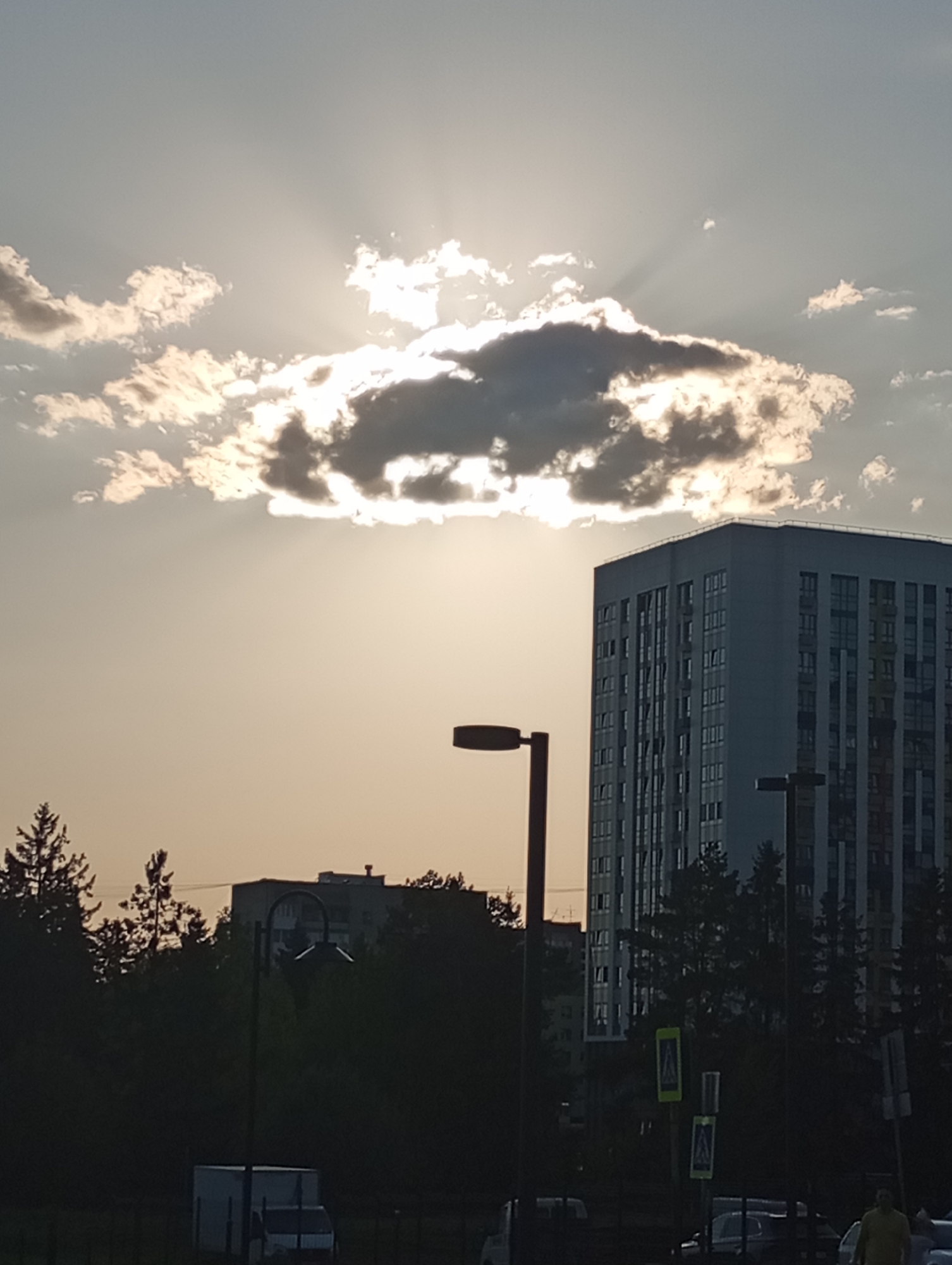 The image shows a view in what appears to be the city of Izhevsk, Russia. A tall, light-colored apartment building with numerous windows is visible in the right half of the frame. To the left of the apartment building is a darker skyline of trees and additional buildings further in the distance. A few streetlights are scattered throughout the scene. In the foreground, there are some vehicles parked and at least one person visible wearing a yellow shirt. The sky is predominantly bright with a few scattered clouds. A prominent cloud formation blocks the sun, creating sunbeams that radiate outwards. The lighting suggests either sunrise or sunset. The presence of a pedestrian crossing sign indicates this is a pedestrian area and likely a street intersection.