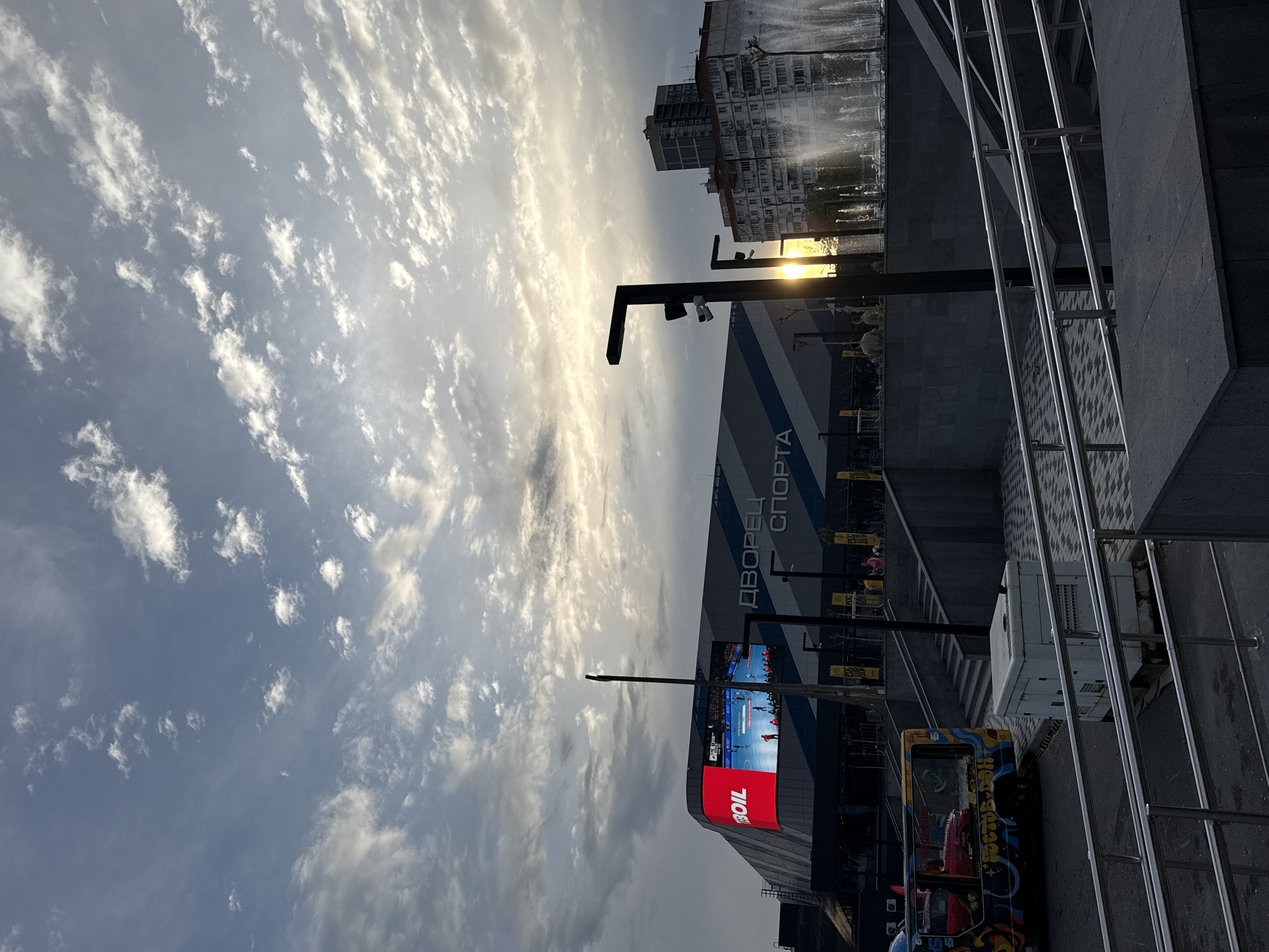 The image shows a view of the Rostov-on-Don Sports Palace in Rostov-on-Don, Russia. The sky is filled with clouds, and the sun is visible behind a lamp post near the Sports Palace. The Palace has a large screen displaying a sports game. In the lower foreground, there are stairs, a painted trolley bus, and metal railings. In the background, a building with multiple windows is visible, along with a fountain.