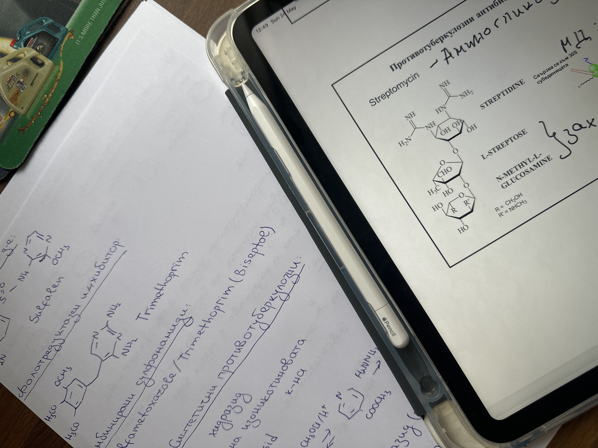 The scene shows a flat surface, likely a desk or table, with several items arranged on it. On the left side of the frame, there's a sheet of white paper filled with handwritten notes in blue ink, appearing to be related to organic chemistry or pharmacology. The notes include chemical structures and names like "Sulfalen," "Trimethoprim," and "fametoxazole."
In the center, there is a white Apple Pencil laying between the paper and an iPad. The iPad is open, displaying a digital document also related to chemistry, specifically the structure of Streptomycin. The iPad shows the time and date as "12:49 Sun 24 May."
A portion of a mousepad featuring motor oil branding is visible in the upper left corner.
The language in the handwritten notes appears to be Bulgarian. This is a likely indication that the location is Sofia, Bulgaria.