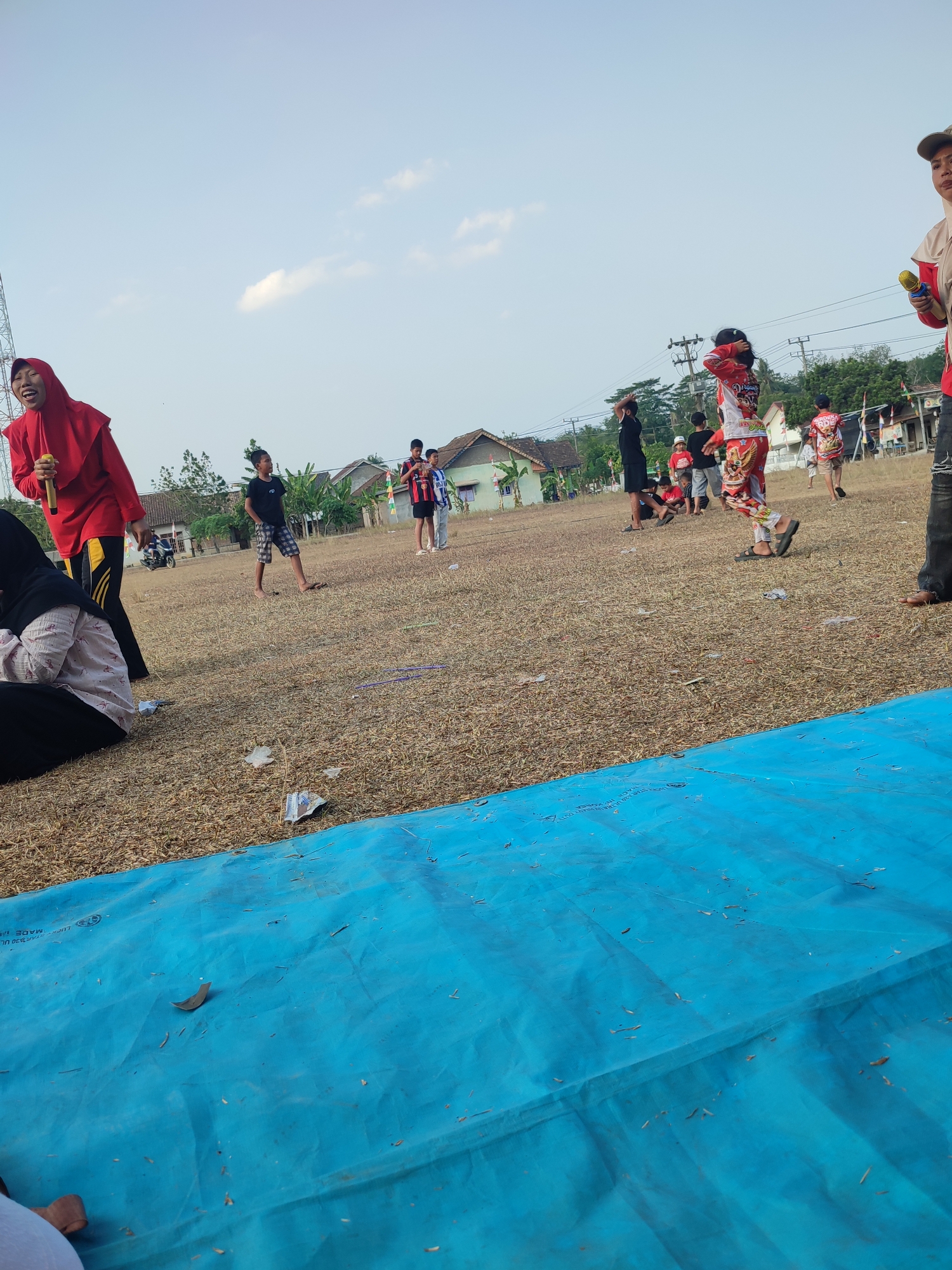 In Nusa Jaya, Indonesia, on a dry, grassy field, a group of people, including children and adults, are gathered outdoors. The sky is clear with a few scattered clouds. In the foreground, a large blue tarp covers a portion of the ground. Several individuals are scattered across the field. On the left, a woman wearing a red hijab and red top is holding a microphone. Further back, a young boy in a black t-shirt and checkered shorts stands. Several other children are visible in the middle ground, some in brightly colored clothing. In the background, houses with tiled roofs are visible, along with trees and utility poles. The overall atmosphere appears to be a casual outdoor gathering or event.