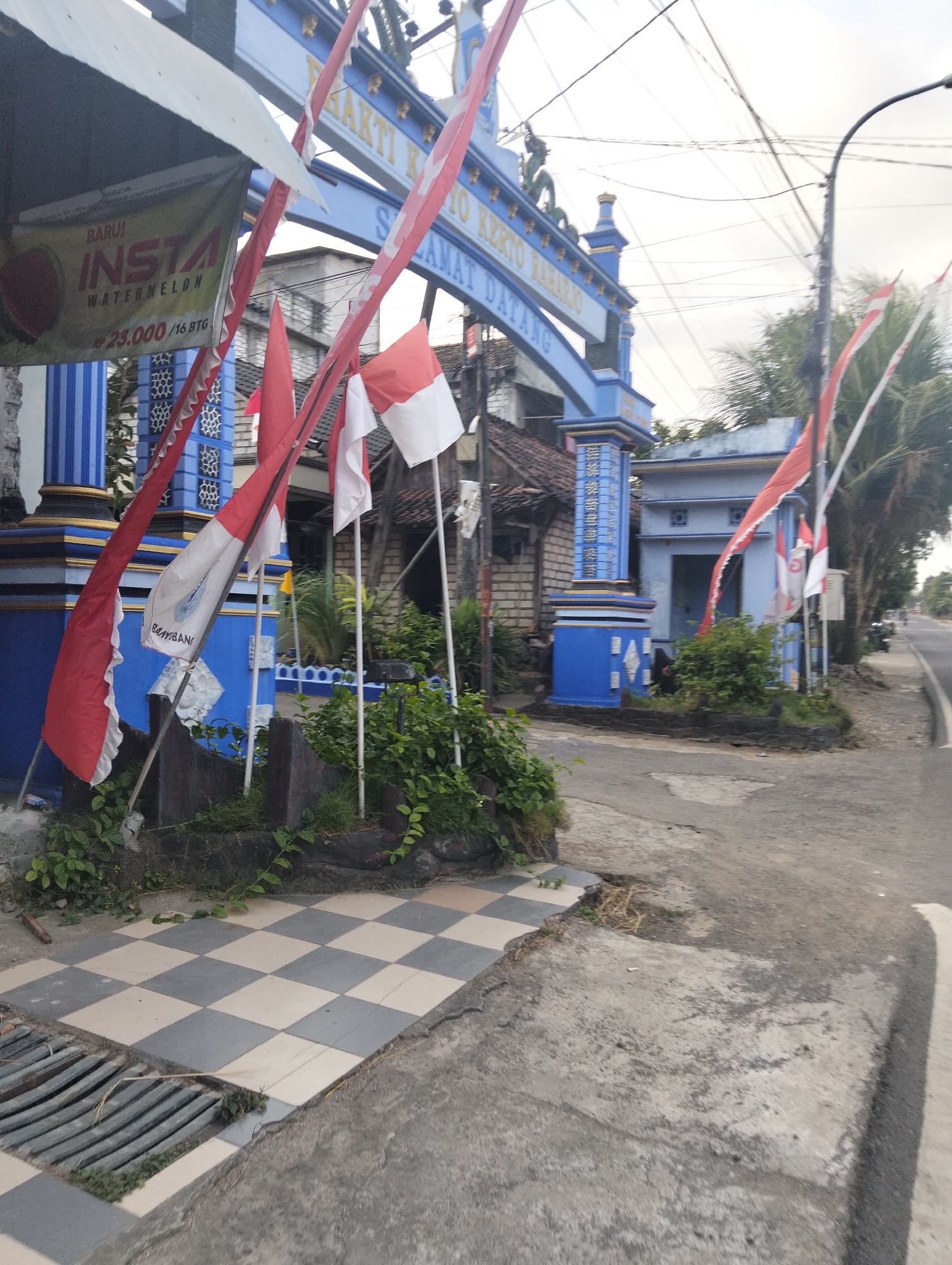 The image captures a street scene in Dagan, Indonesia, focusing on a blue archway adorned with Indonesian flags and decorative elements. The archway has text that reads "BHAKTI KEPADA RAHAYU" and "SELAMAT DATANG." To the left of the archway, a banner is visible with the text "BARU! INSTA WATERMELON" and a price of "23.000 /16 BTG." The ground in the foreground features a checkered tiled pathway adjacent to a concrete road. Various red and white flags are prominently displayed on poles, some fluttering in the breeze. Green foliage and bushes are present around the base of the archway and along the pathway. In the background, there are buildings and trees, and the sky is overcast. The overall impression is one of a welcoming gateway, possibly marking an entrance to a village or a significant location within Dagan.