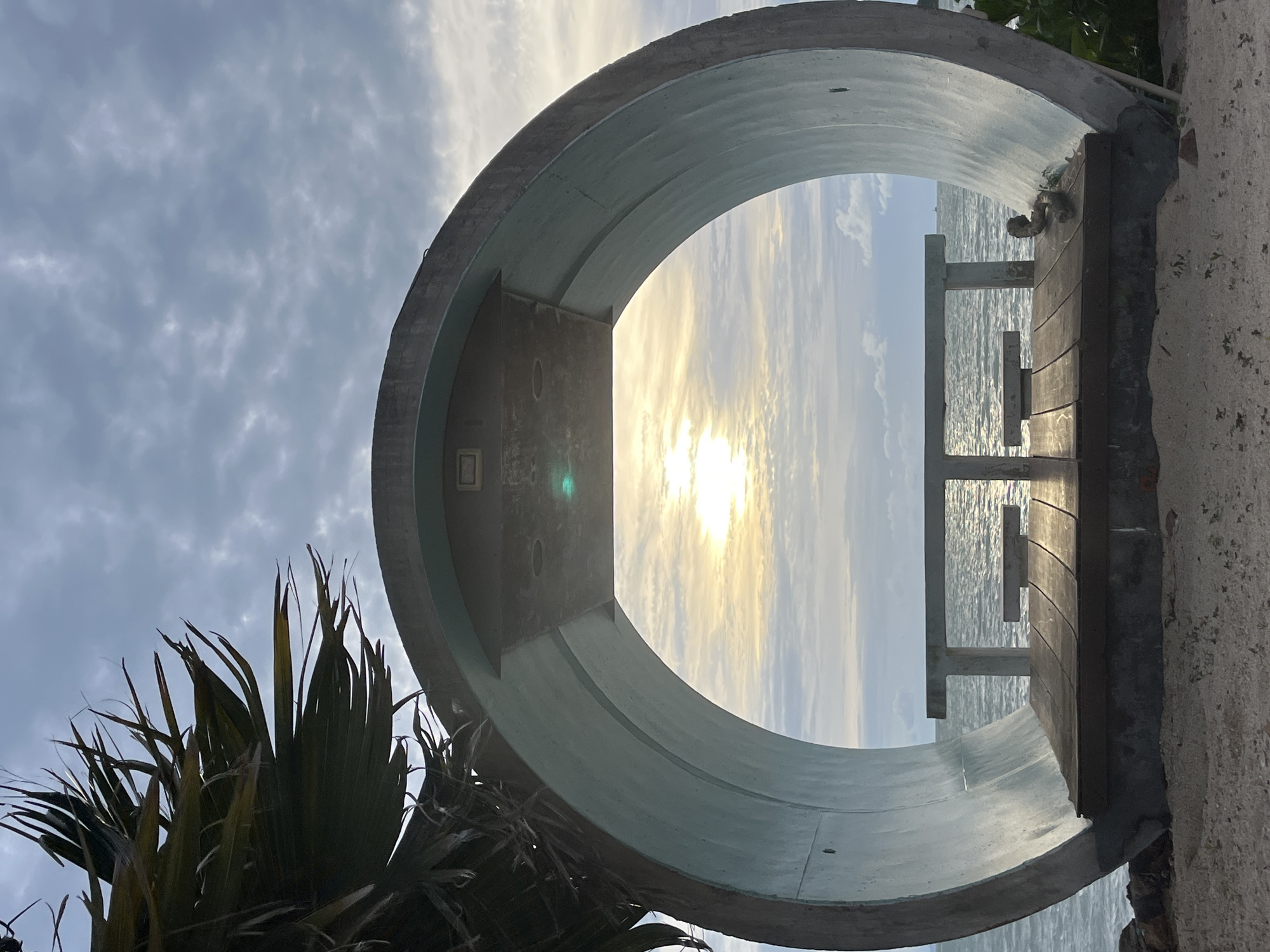 The image depicts an outdoor seating area with a circular concrete structure framing a view of the ocean and sky. The circular structure, painted a light blue-grey, is open at the front, revealing a wooden platform with two small concrete benches and a concrete table. Beyond the platform, the sea stretches out under a cloudy sky, with the sun partially visible through a break in the clouds, casting a warm glow on the water. The foreground consists of sandy ground with sparse vegetation. To the left, a large palm frond hangs over the edge of the circular structure. The overall scene suggests a tranquil spot on the coast of Ko Samui District, Thailand, likely intended for relaxation and enjoying the seascape.