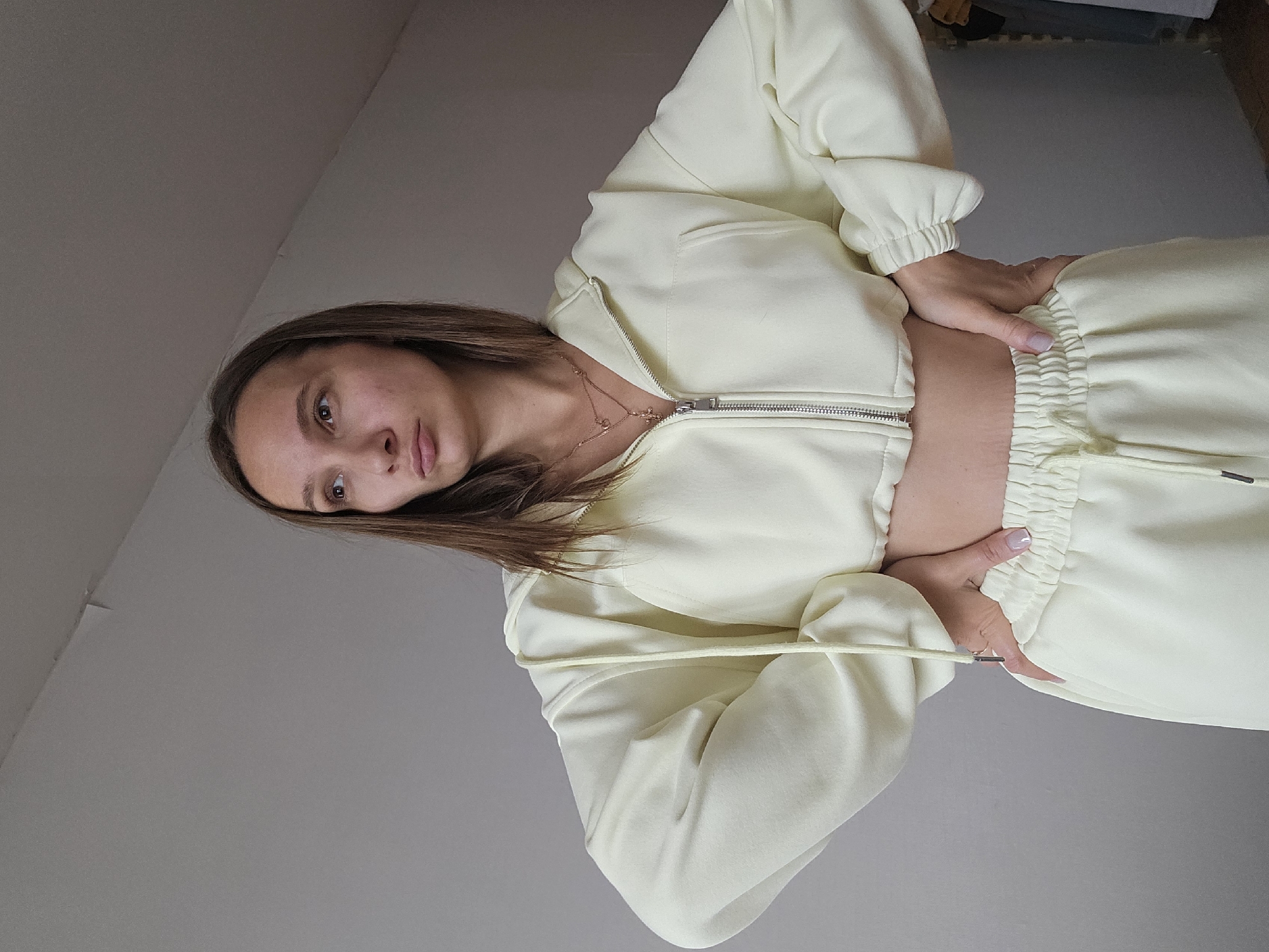 A young woman with light brown hair and a light complexion stands with her hands on her hips, looking off to the left. She is wearing a pale yellow cropped zip-up hoodie and matching sweatpants. The background is a plain, light gray wall with a slight upward slope towards the ceiling. There is a subtle shadow on the wall behind her. The lighting is soft and diffused. The location is not specified in the image.