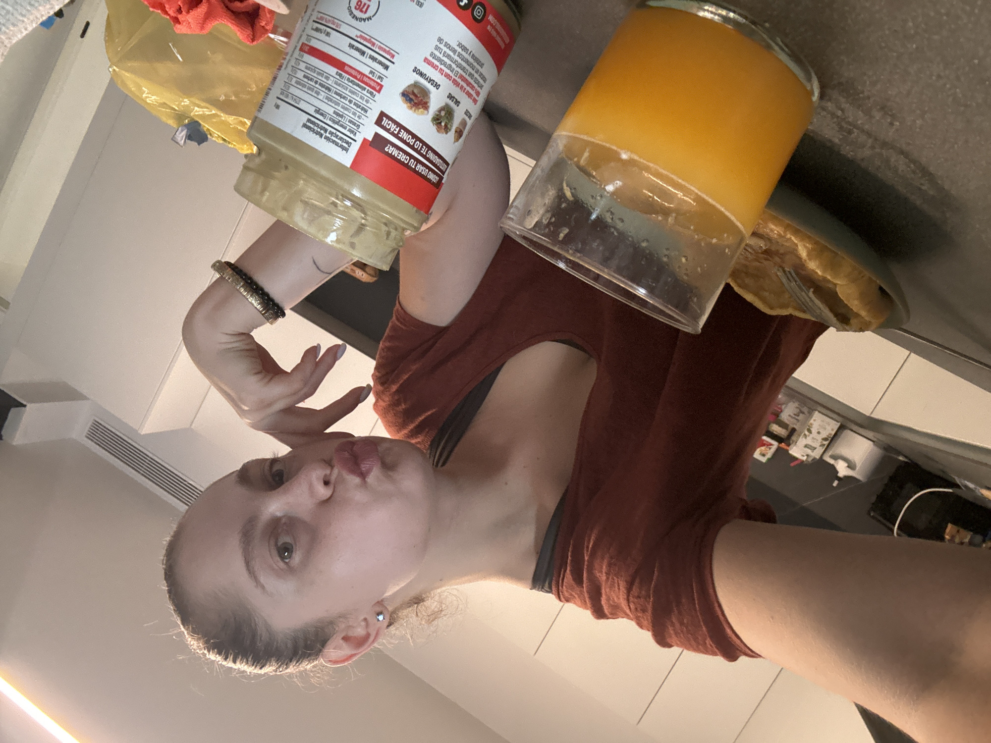 A young Caucasian woman with her hair pulled back in a bun and wearing a rust-colored t-shirt and a grey sports bra is taking a selfie in a kitchen. She is puckering her lips and resting her chin on her hand, which is adorned with a bracelet. To her left, a glass of orange juice and a plate of what appears to be a pastry are on a dark grey countertop. In the foreground, a jar of tahini is visible with its nutritional information label facing the camera. The kitchen cabinets are white, and the lighting suggests it is daytime. The setting appears to be a residential kitchen.