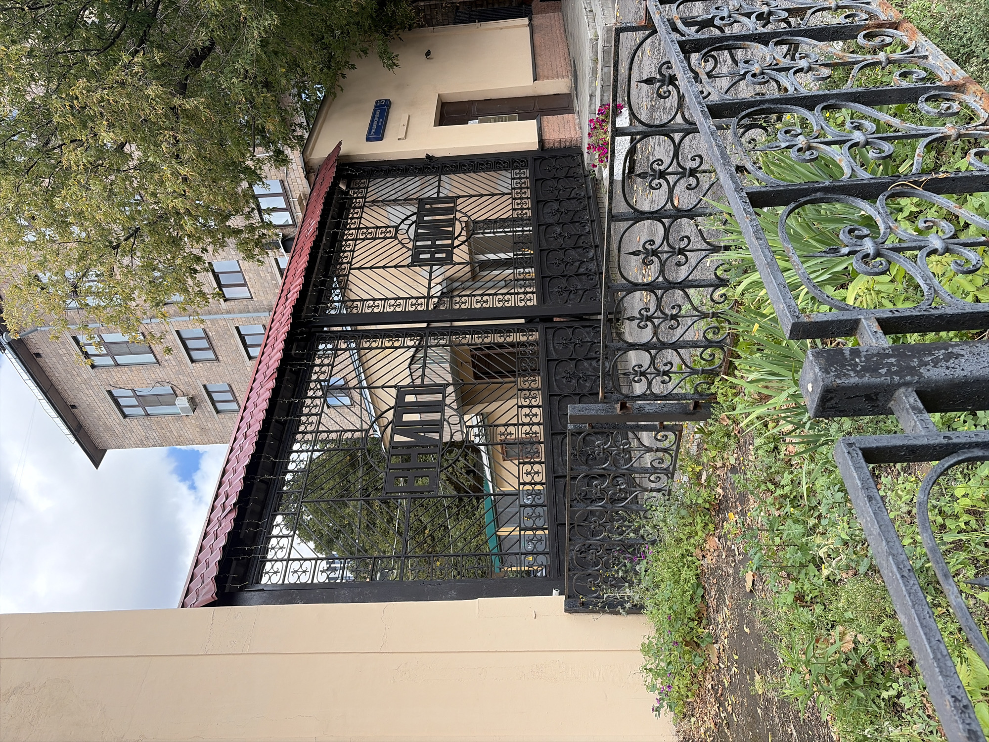 The image shows a black ornate metal gate with a red tiled roof overhead, set against a pale yellow building on the right and a light beige building on the left. The gate has a section with the Cyrillic letters "ВНИИ" (VNIY) visible. Behind the gate, there are more buildings and trees. A street sign in blue with white text is visible on the yellow building, indicating "2-я МУЗЫКАЛЬНЫЙ ПЕРЕУЛОК 15" (2nd Musical Lane 15), which is a location in Moscow, Russia. The foreground features a black metal fence with decorative elements and overgrown greenery with some scattered leaves. The sky is cloudy. There are no people visible in the image.