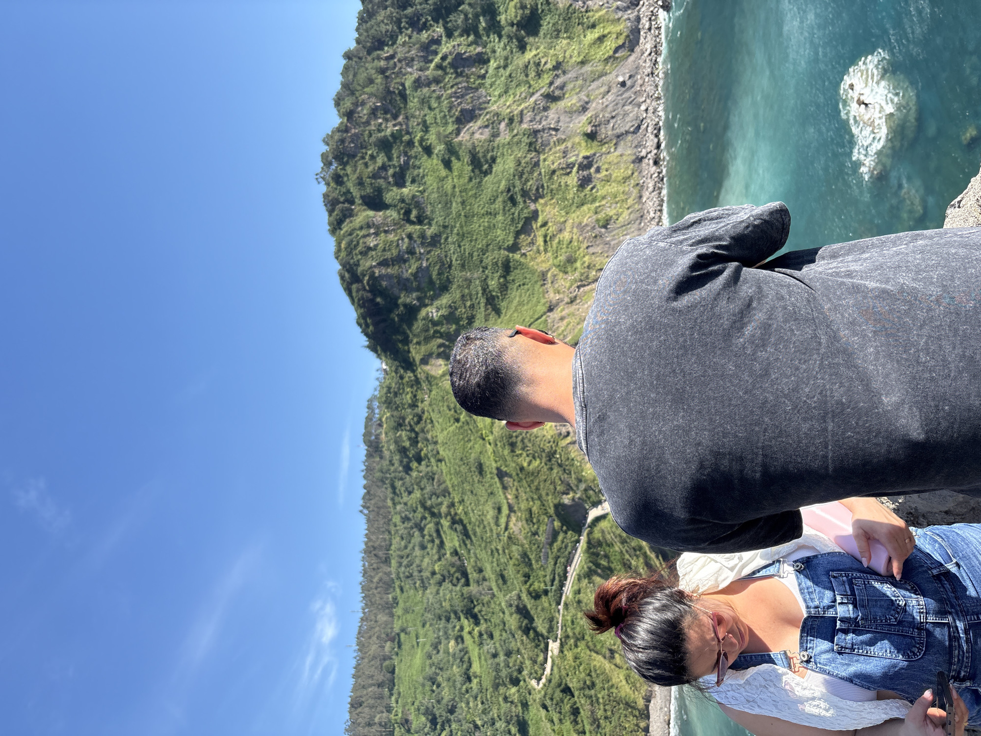 Two individuals stand on a cliff overlooking the ocean. The person in the foreground, facing away from the camera, wears a dark-colored t-shirt. To their left, a person wearing denim overalls and a white top looks down at their phone and a pink object they are holding. The background features a vibrant green, treed hillside with a winding path visible on its slope, leading down towards the turquoise ocean. Rocky outcrops are present in the water. The sky is a clear, bright blue. The location is identified as Bakio, Spain.