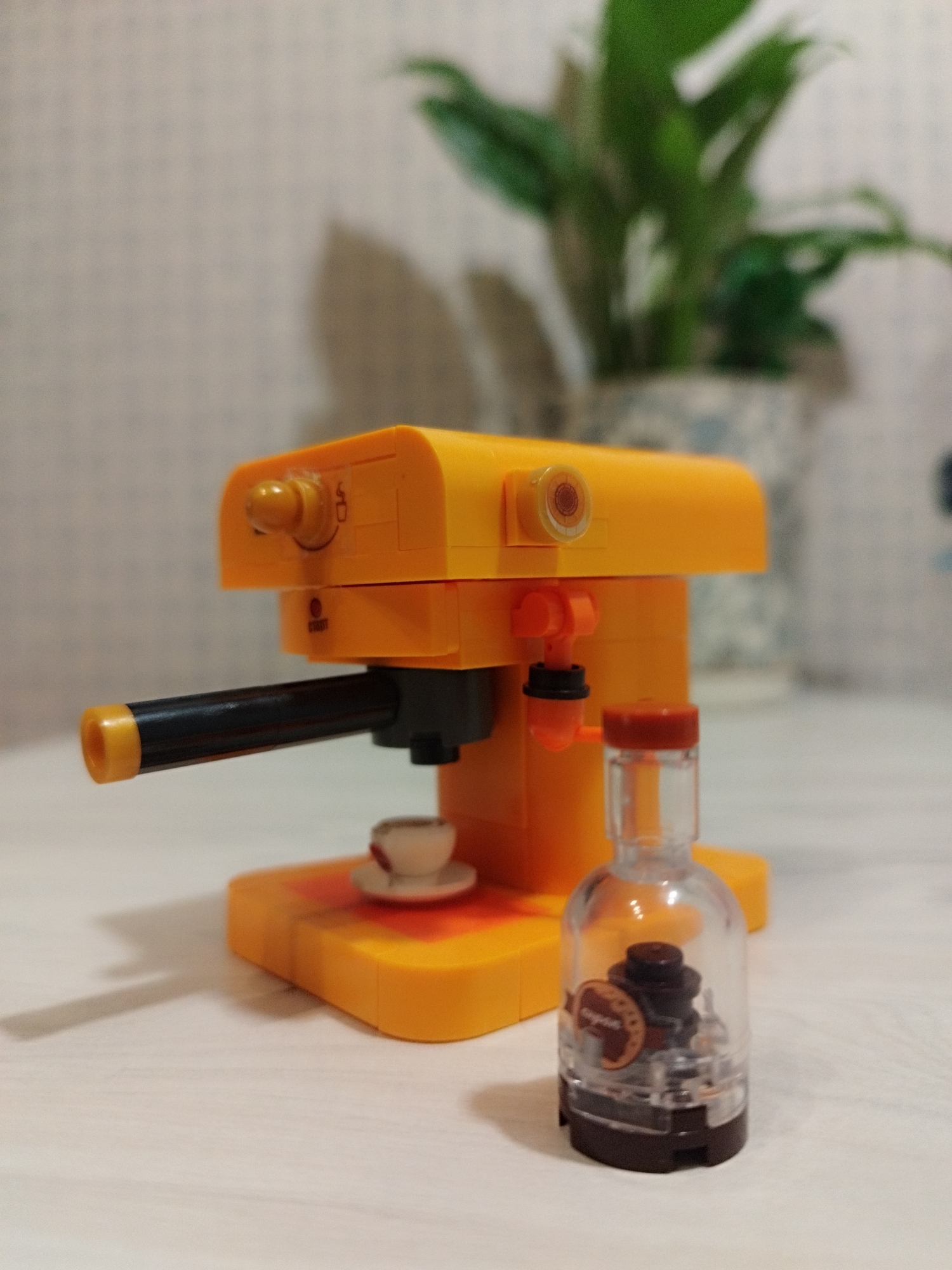 A close-up, eye-level shot shows a miniature yellow toy espresso machine with a black portafilter. A small, white teacup and saucer sit on the machine's base. To the right of the machine is a clear plastic bottle with a brown cap, containing a dark brown substance and a circular label with a picture of a coffee maker. In the background, a blurred green plant is visible in a potted planter against a textured, light-colored wall. The scene appears to be indoors, on a white, wooden surface. The location is not in Saint Petersburg, Russia.