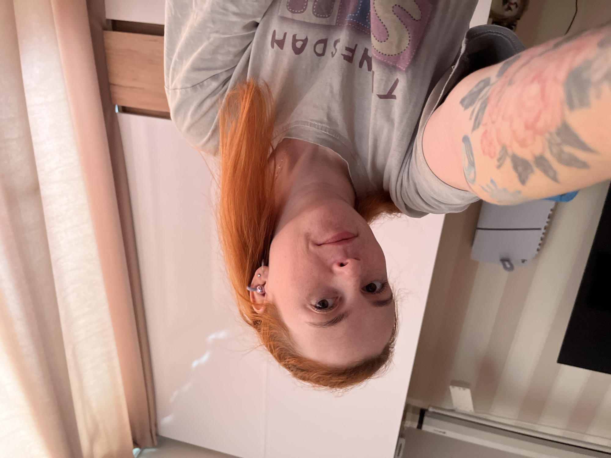 The image shows a person with red hair, facing the camera. The person is wearing a light-colored t-shirt with text on it. The t-shirt has a graphic design and appears to have the words "TUESDAY" printed on it in a retro font. The person has a tattoo on their right arm, featuring floral imagery in shades of pink and gray. The background is blurry but suggests an indoor setting, possibly a room with curtains and white furniture. The lighting indicates it might be daytime, with natural light filtering in. The person's expression is neutral, and the overall mood is casual and relaxed. The image is rotated, with the person's head at the bottom and their feet at the top. There are no obvious external location cues that would specifically link it to Chaikovsky, Russia.