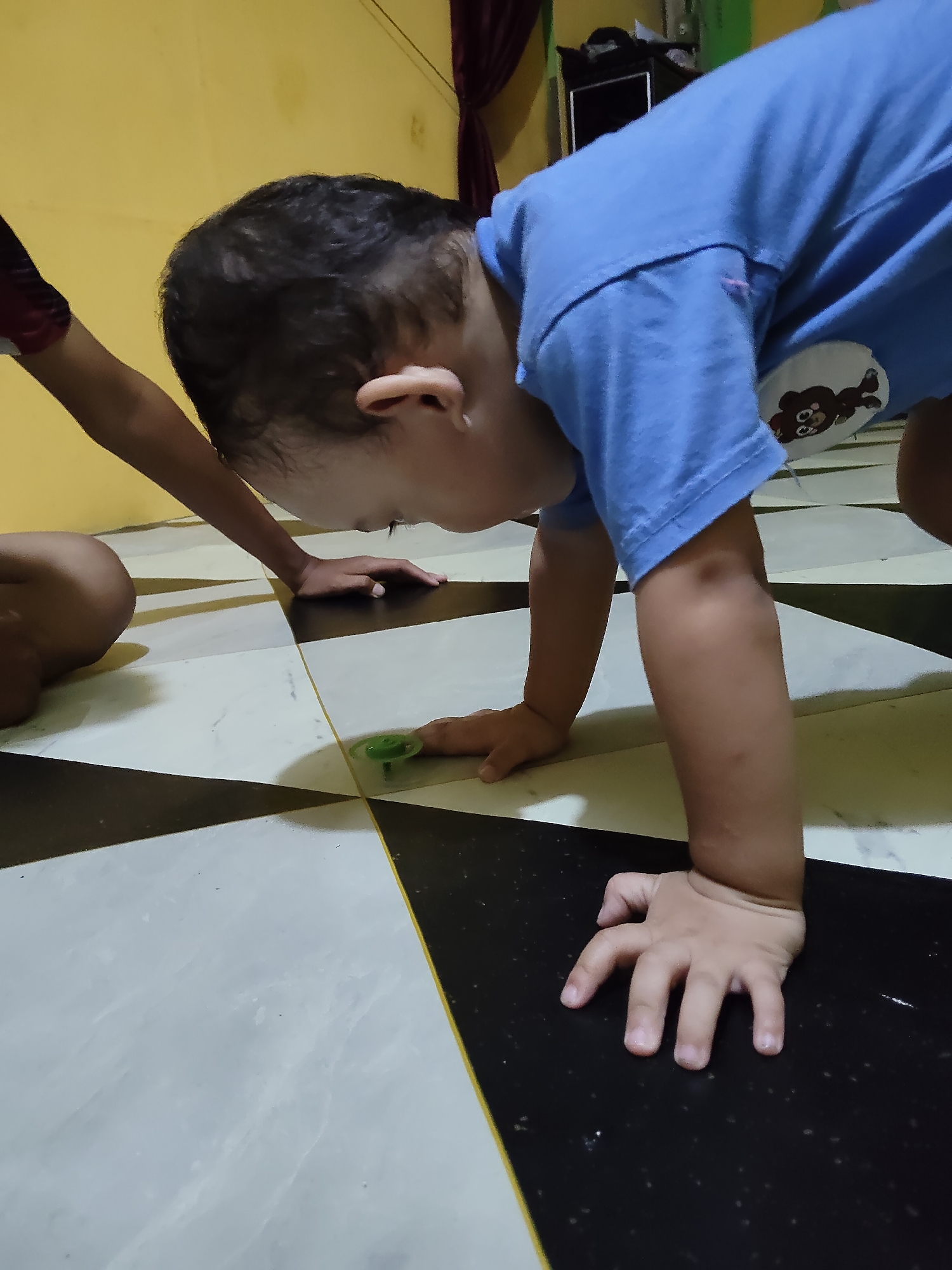 In Tanah Grogot, Indonesia, a young child is on all fours on a black and white tiled floor. The child, wearing a blue t-shirt with a bear graphic, is looking down at a small green spinning top on the floor. Another child's arm, wearing a red and black shirt, is visible to the left of the frame. The background consists of a yellow wall and dark curtains.