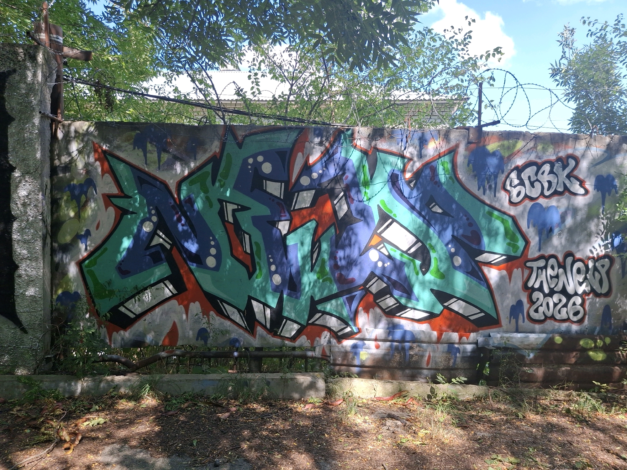 An exterior, eye-level, full shot shows a concrete wall covered in graffiti on a sunny day. The wall is topped with barbed wire and surrounded by trees. The graffiti features large, stylized letters in shades of blue, green, and orange, with white highlights. To the right of the main graffiti, the words "SCBK" are spray-painted in white with a black outline, followed by "THE NEW 2026" in a similar style. The ground in front of the wall is covered in dry leaves and dirt, with sparse green foliage growing along the base of the wall.