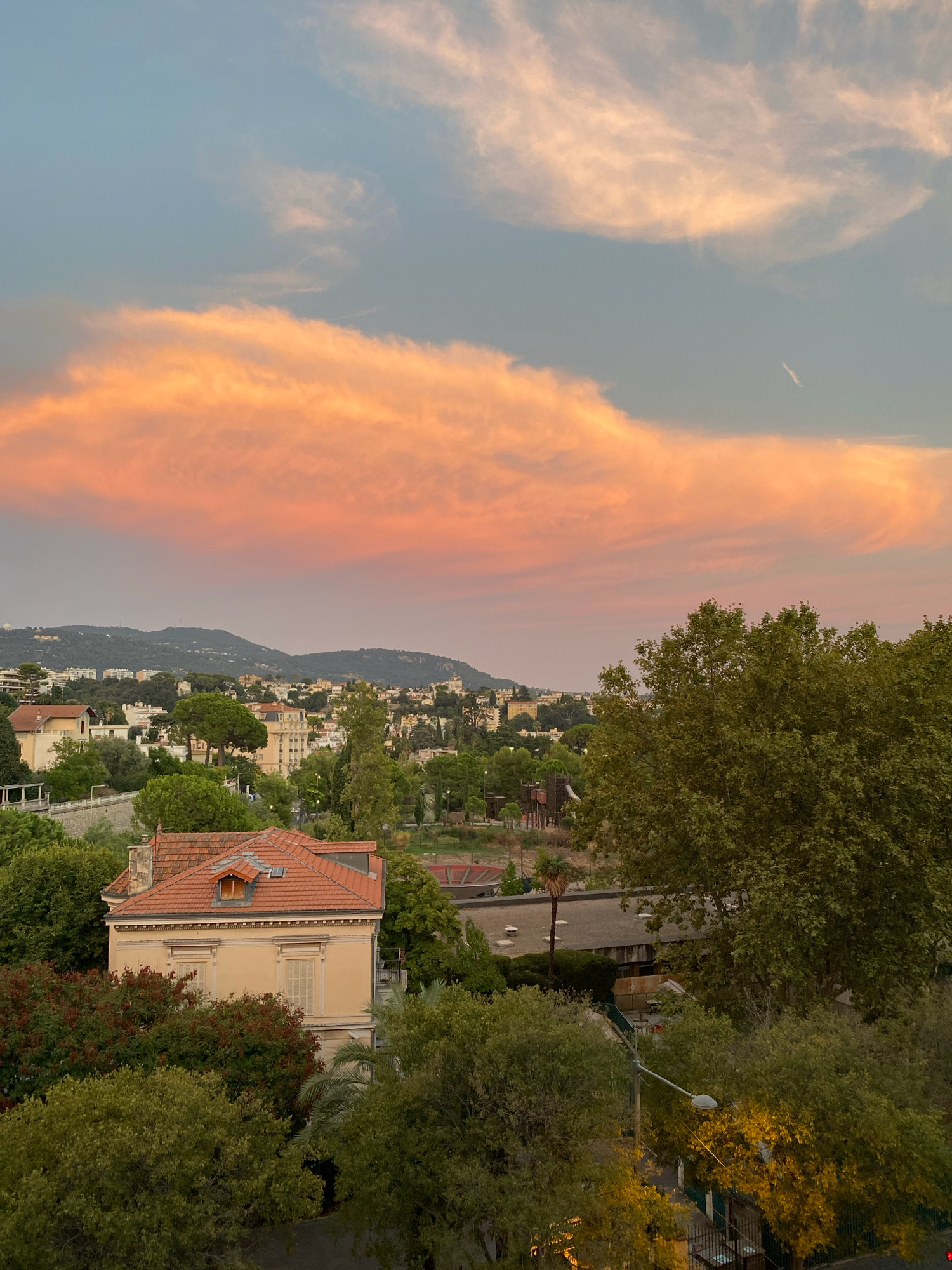 This is a medium-range, aerial view of a suburban area in Nice, France, captured at dusk. The sky is dominated by a dramatic band of clouds tinged with orange and pink from the setting sun, contrasting with the softer blue of the upper atmosphere. Below, a dense collection of buildings, many with red-tiled roofs, are nestled amongst rolling green hills. Lush trees, a mix of deciduous and palm varieties, are abundant throughout the landscape, some showing the golden glow of the evening light. In the foreground, a charming, light-beige house with a red-tiled roof and two dormer windows is partially obscured by trees. A streetlamp with its light illuminating nearby foliage is also visible in the lower right corner. The scene evokes a peaceful, end-of-day atmosphere in a picturesque French city.