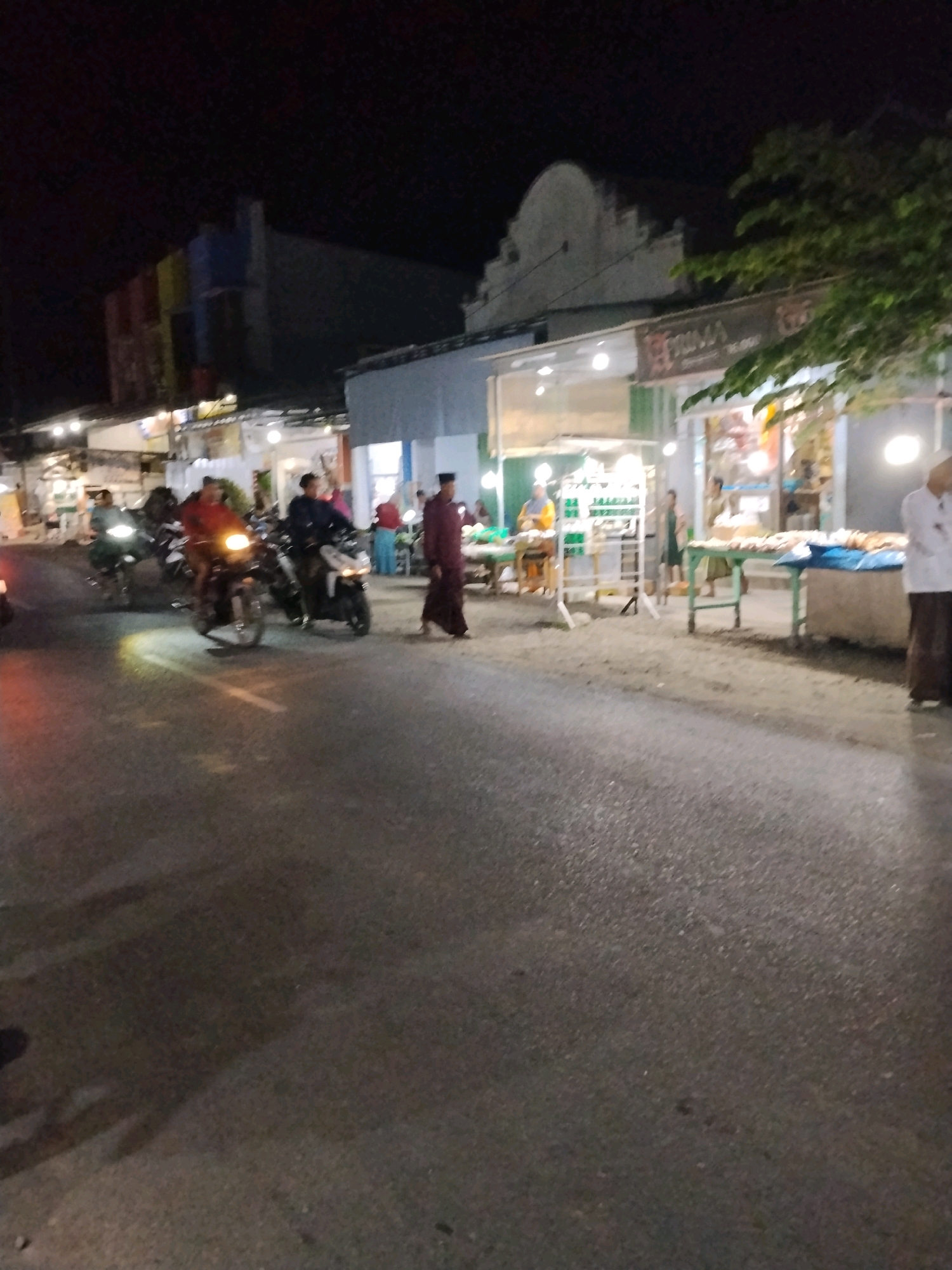 The image depicts a street scene at night in Panggungan, Indonesia, likely near a marketplace or commercial area. Several individuals are present, with a group of people on motorcycles in the foreground. One person is walking in the middle ground, wearing a traditional maroon garment and a cap. Further back, individuals are seen interacting around stalls or shops, with some appearing to be vendors and customers. The environment includes buildings with illuminated windows and signs, as well as trees and the street itself, which is paved. The lighting suggests artificial illumination from streetlights and shop fronts.