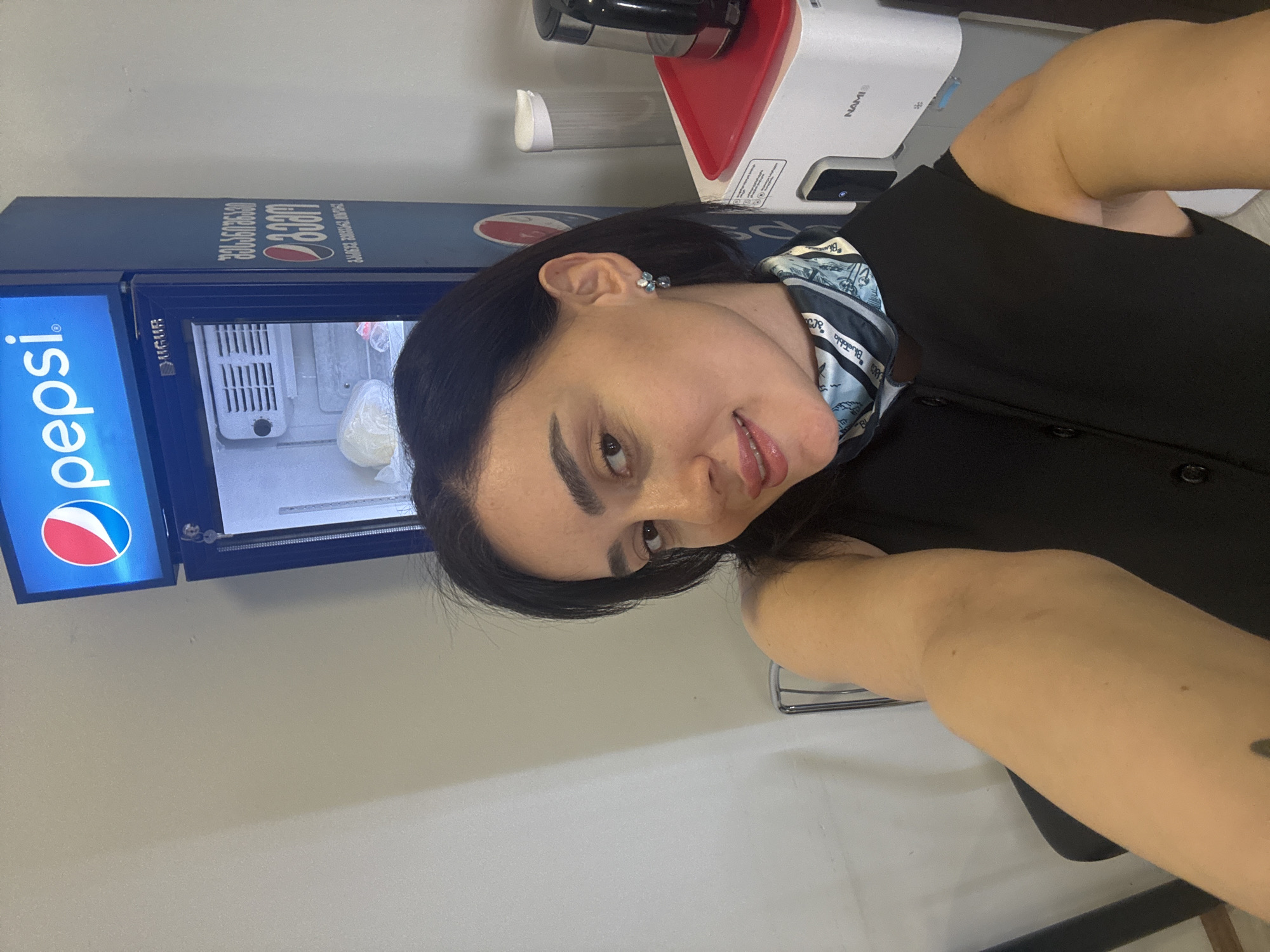 A woman with dark hair, fair skin, and wearing a black top and a blue patterned scarf is posing for a selfie indoors. Behind her is a small, blue Pepsi-branded refrigerator containing items wrapped in plastic. To the right of the refrigerator is a white water dispenser with red accents. The wall behind the woman and the refrigerator is a light neutral color. The location appears to be a business or commercial setting. The presence of Pepsi branding and a Georgian phone number (595395160) suggests the image was taken in Tbilisi, Georgia.