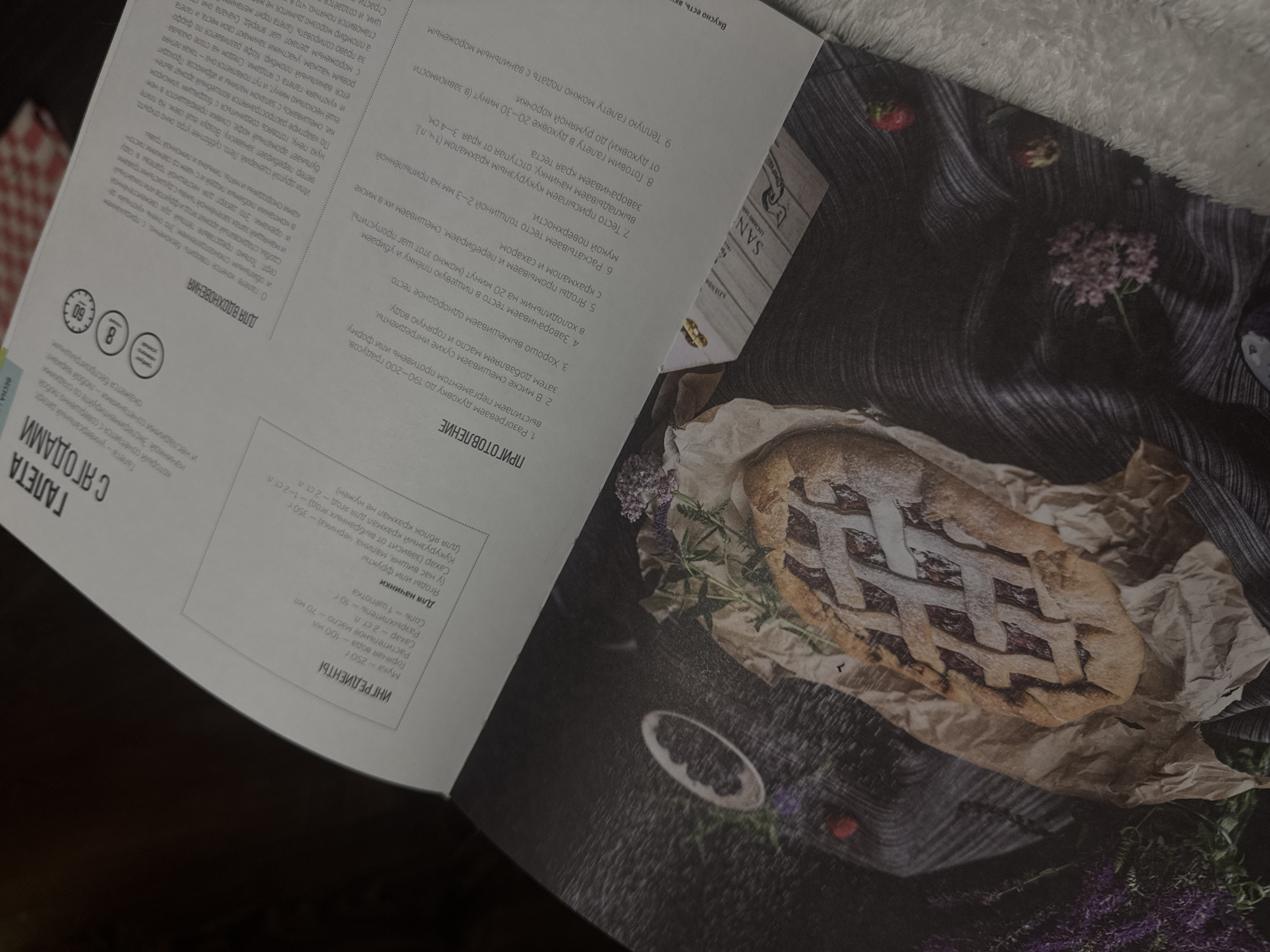 Here's a detailed description of the image:
**General Description:**
The image is a photograph of an open magazine or cookbook spread. On the right-hand page, there is a picture of a berry galette, a rustic fruit tart. The left-hand page contains text, presumably a recipe or description of the galette.
**Specific Details:**
* **Galette:** The galette is placed on a dark surface with a wrinkled piece of parchment paper underneath. It has a lattice crust and appears to be filled with a dark berry filling. There are scattered purple flowers and what looks like some berries around the galette, likely for decorative purposes.
* **Magazine Text:** The text is in Russian. The title of the recipe appears to be "Galeta S Yagodami," which translates to "Galette with Berries." It includes a list of ingredients and instructions.
* **Surroundings:** The image is taken at a low angle, which gives the impression the viewer is looking down on the page. There is a dark, possibly wooden, background with some decorative elements.
**Environment and Mood:**
The lighting is soft and slightly muted, giving the image a warm, cozy feeling. The overall impression is that of a simple, rustic dessert recipe in a lifestyle or food magazine.
**Location:**
There is no indication from this image that the image was taken at Izhevsk, Russia.