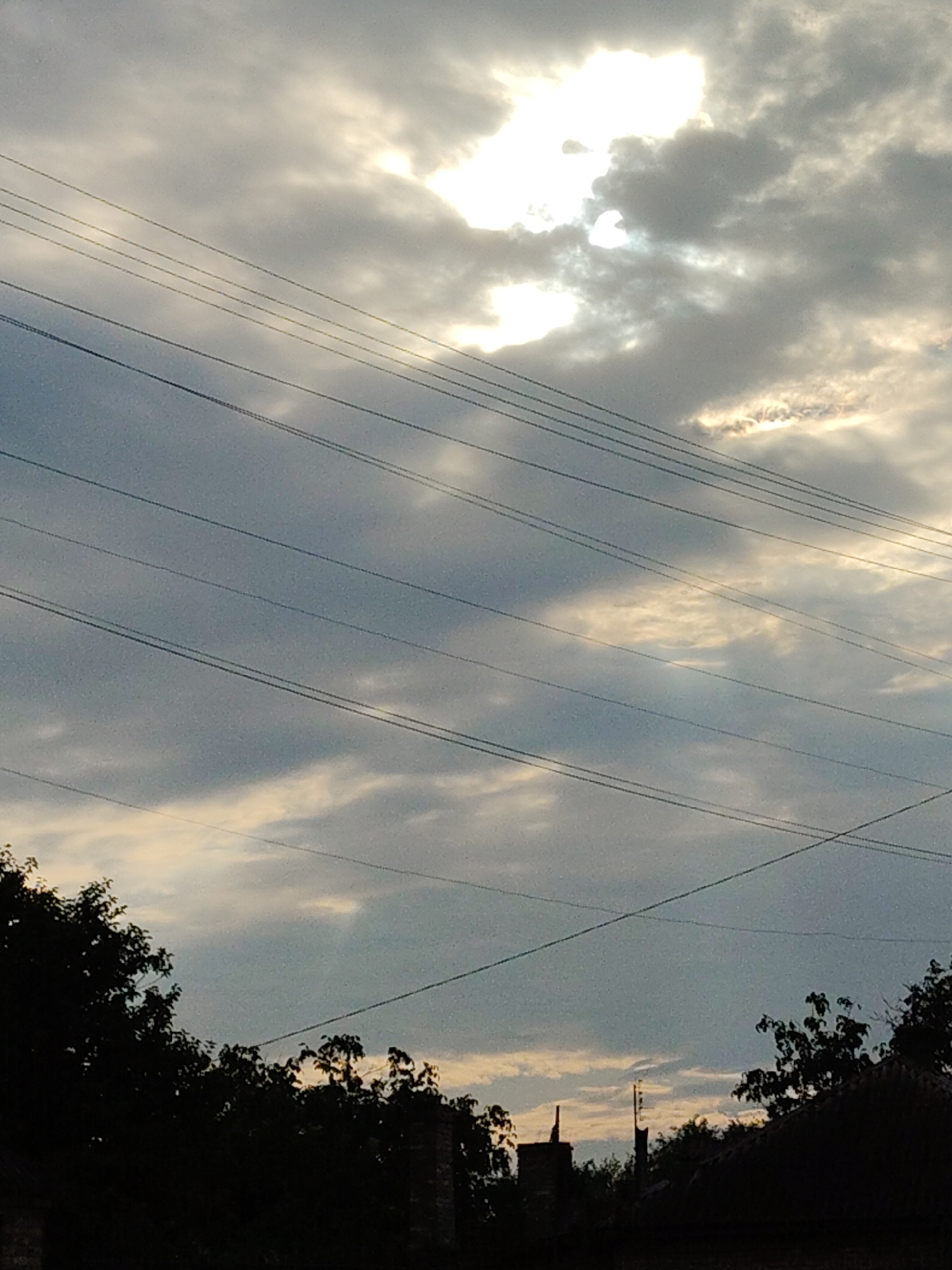 The image depicts a cloudy sky over Cherkasy, Ukraine, with multiple power lines stretching horizontally across the frame. The sun is partially obscured by clouds, creating a bright glow in the upper portion of the sky. Silhouetted trees and the tops of buildings are visible along the bottom edge of the image. The scene appears to be an outdoor view, likely from a residential area or a vantage point looking towards the sky. There are no people or discernible actions in the image.