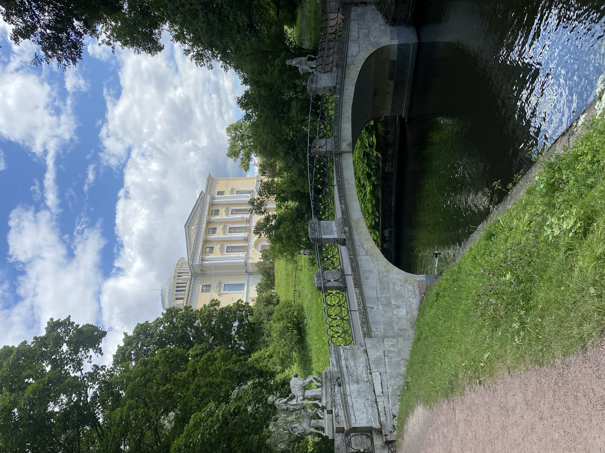 The image depicts a stone bridge spanning a dark body of water, with a neoclassical palace in the background. The bridge, adorned with ornate black metal railings and classical sculptures at its ends, leads towards a grand yellow palace situated on a grassy hill. Lush green trees frame the scene, and a dirt path runs along the left side of the water. The sky above is a mix of blue with scattered white clouds. This view captures the essence of the Pavlovsk Park, a suburban imperial park located in the town of Pavlovsk, near Saint Petersburg, Russia, renowned for its picturesque landscapes and classical architecture.