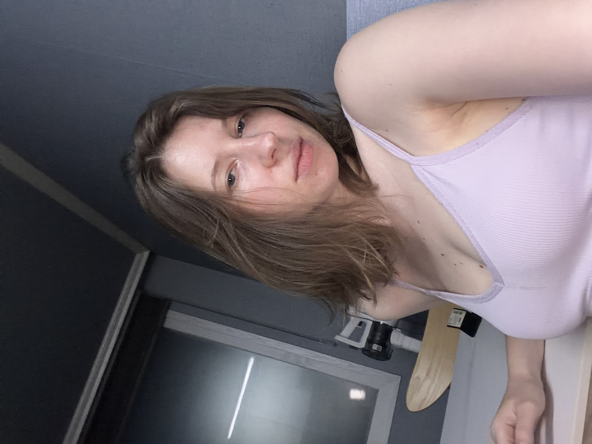 This is a selfie taken indoors, likely in a residential setting in Dongducheon-si, South Korea. The subject is a young Caucasian woman with shoulder-length brown hair and a light complexion. She is wearing a light purple ribbed tank top. Her gaze is directed slightly off-camera, and her expression is neutral. The background is predominantly a gray wall. To the left of the subject, a window with a reflective surface is visible, and behind her, a light-colored wooden object, possibly a skateboard, is leaning against the wall. A black cylindrical object, which could be a camera or lens, is also visible near the window. The lighting appears to be artificial and somewhat dim.
