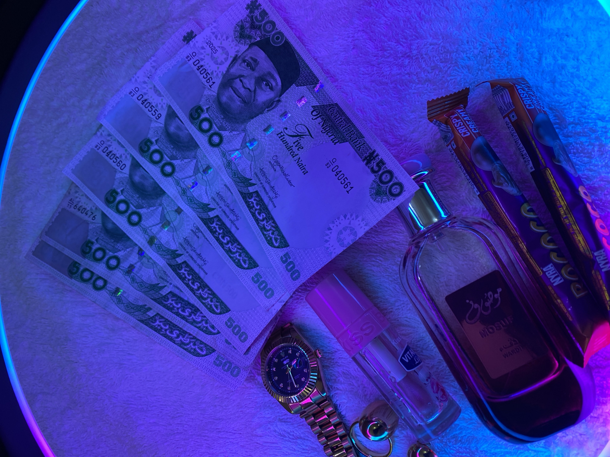 This media file presents a flat lay arrangement of various items on a white, fluffy textured surface, brightly illuminated by blue and purple artificial lighting, hinting at an indoor setting in Maigizo, Nigeria. A glowing arc in the upper left suggests the light source might be an LED ring light.
The central subjects are several Nigerian 500 Naira banknotes, fanned out diagonally. Visible text on these notes includes "Central Bank Of Nigeria," "Five Hundred Naira," "Dr. Nnamdi Azikiwe," and the Arabic script "خمسمئة نيرة" (khamsumi'ah nayra). Serial numbers such as "040561," "040559," "040560," and "640476" are legible, along with the year "2025" and the signatures of the Governor (Olayemi Michael Cardoso) and Director of Currency Operations.
Accompanying the currency are personal accessories and snacks. A gold-toned wrist watch, resembling a Rolex Oyster Perpetual Datejust with a diamond-set dial and fluted bezel, is prominently displayed. Nearby, a clear bottle of lip gloss or lip balm is visible, labeled "KISS" and "VITAL T." A larger, elegant glass perfume bottle, featuring Arabic script "موضوون" (MOSUF) and "WARDI" on its label, stands upright. Two "NYRA CRISPY CREAMY" chocolate bars are positioned on the right, showing partial text like a barcode number "8906121072655" and a phone number.
No specific activity or event is depicted, but the arrangement conveys an aesthetic display of personal luxury items and everyday treats. The striking artificial lighting creates a modern and vibrant atmosphere.