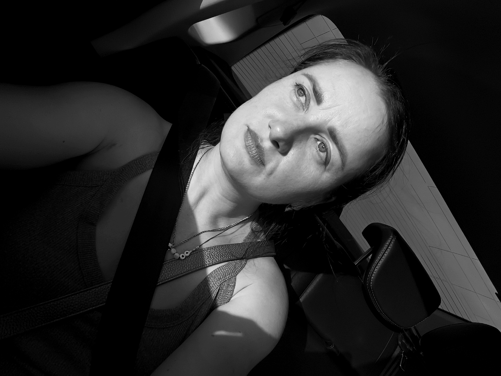 A black and white, high-angle, close-up selfie shows a woman wearing a seatbelt inside a vehicle. The woman is looking upwards with her head tilted to the left. She has dark hair, fair skin, and is wearing a sleeveless top and a necklace. The background is dark, with some visible interior car elements such as a headrest and what appears to be a window with a grid pattern. The lighting suggests it is daytime, with shadows cast across the woman's face and upper body. There is no indication of the specific location within Lelystad, Netherlands.