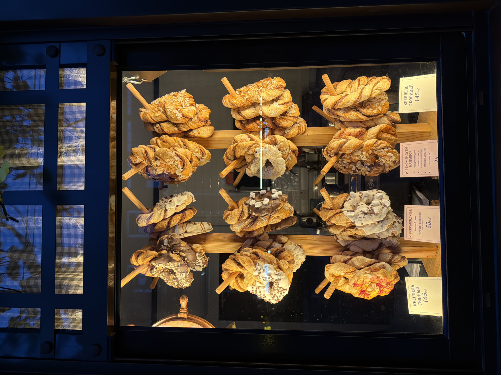 The image shows a display window of a bakery in Svetlogorsk, Russia. The window features a variety of twisted pastries, likely pretzels or sweet buns, hanging from wooden sticks. The pastries are arranged in multiple tiers. Some pastries are topped with chocolate, nuts, white icing, or marshmallows. Below the pastries, there are price tags in Russian. One tag reads "КРЕНДЕЛЬ СЫРНЫЙ 165 руб." (Cheesy Pretzel 165 rubles). Another tag says "РЕКОМЕНДУЕМ НА ВЫБОР" (Recommended to choose from) with items like "ФУНДУК/МИДАЛЬ" (Hazelnut/Almond), "ШАРИКИ КАРАМ" (Caramel balls), and "КАРАМЕЛЬ СОЛЕНАЯ" (Salted Caramel) priced at 25 rubles, and "МАРШМЕЛОУ" (Marshmallows). A third tag displays "РЕКОМЕНДУЕМ С ГОРЯЧИЙ ШОКОЛАД (БЕЛЫЙ/ТЕМНЫЙ) 55 руб." (Recommended with hot chocolate (white/dark) 55 rubles). A fourth tag shows "КРЕНДЕЛЬ С КОРИЦЕЙ 145 руб." (Cinnamon Pretzel 145 rubles). The background of the window is dark, with reflections of patterned fabric and lights visible, suggesting the interior of the bakery or the street outside.