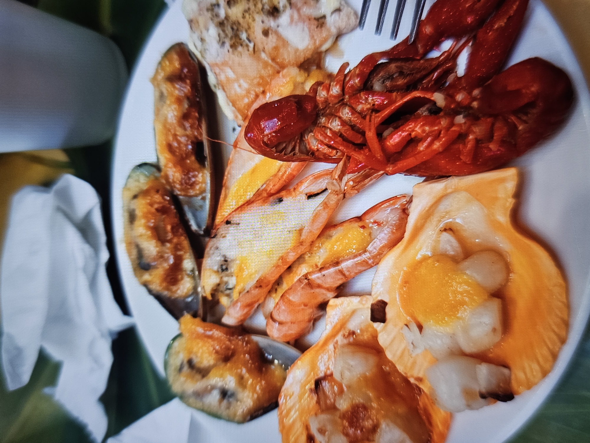A close-up, overhead shot showcases a white plate filled with various cooked seafood dishes. In the foreground, a bright red crawfish lies on its back, its antennae and legs splayed. To the left of the crawfish, two large scallops are served in their shells, topped with a golden-orange sauce. Behind the crawfish and scallops are several grilled shrimp, their shells peeled and bodies curled. Further back, a cluster of mussels in their dark shells are visible, some appearing baked or broiled with a reddish-brown topping. The plate is set against a blurred green surface, and a white paper napkin is visible in the upper left corner. A white cup or container is also partially visible in the upper right. The image does not provide any identifiable landmarks or context that would allow for determination of its location in Amarillo, United States of America.
