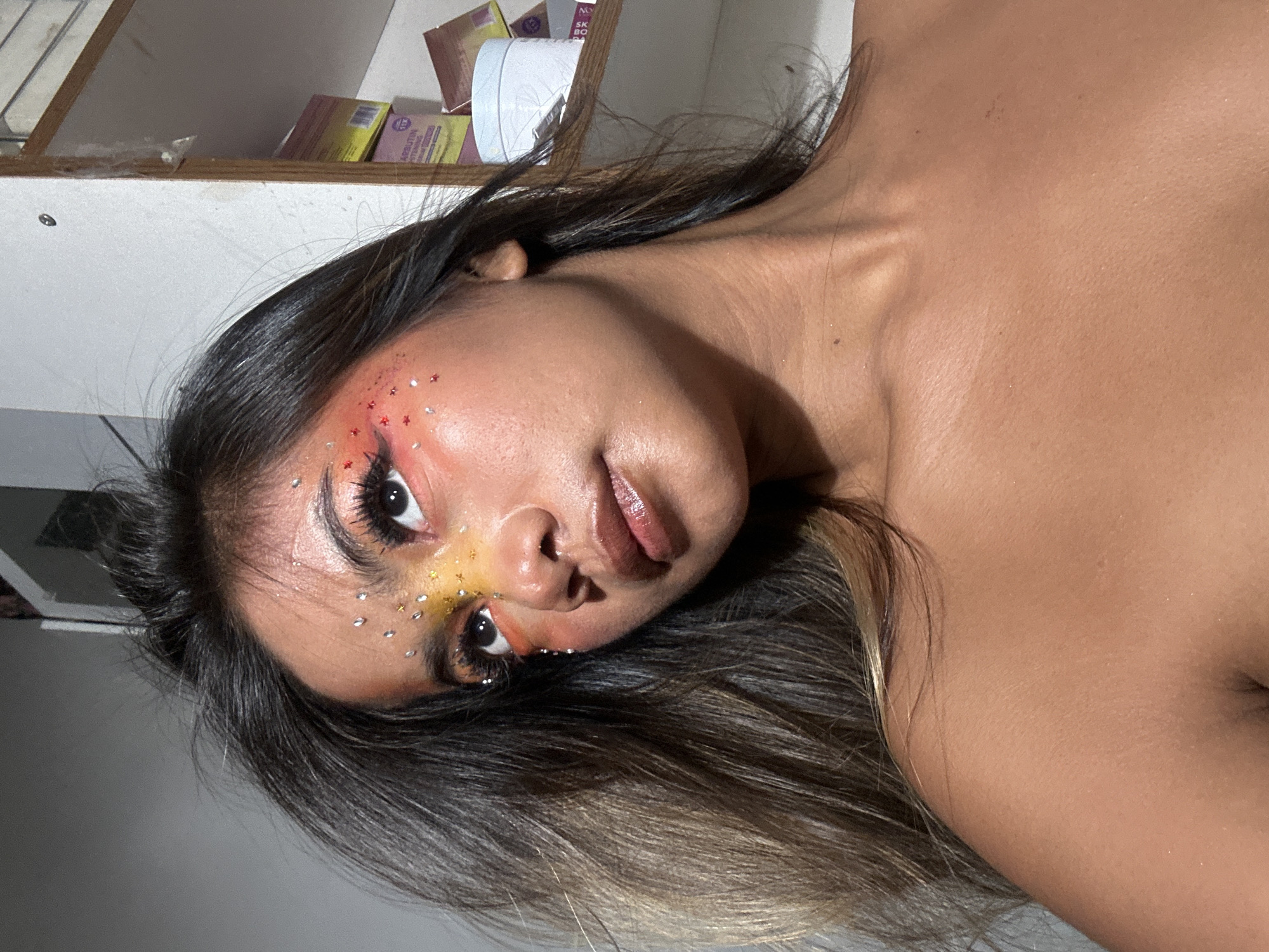 A close-up, slightly angled shot depicts a woman with fair skin and dark brown, ombre hair. She has elaborate makeup with red and yellow eyeshadow and small rhinestones applied around her eyes. Her lips are glossy. The background is blurred, but a white shelf with various boxes is visible. It is impossible to determine if the picture was taken in North Jakarta, Indonesia.
