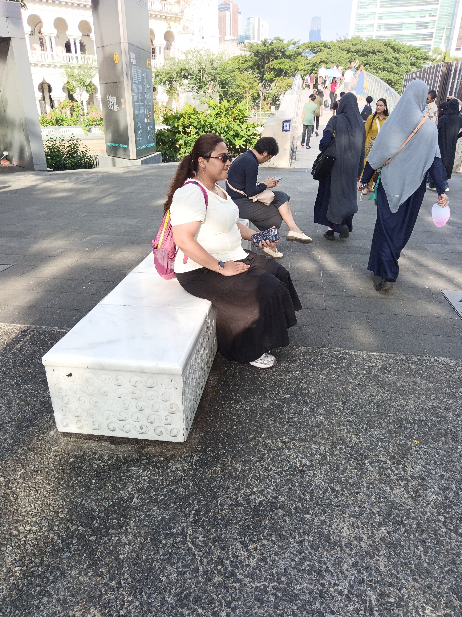 In Kuala Lumpur, Malaysia, at the foot of the Merdeka Square Arch, a woman sits on a decorative white marble bench. She is wearing a white short-sleeved top, a long black skirt, and white sneakers. A pink backpack with yellow straps is slung over her shoulder. She wears sunglasses and her hair is pulled back in a ponytail. Her left hand rests on her lap, and her right hand holds a mobile phone. She appears to be looking at the phone. The bench is adorned with intricate swirling patterns on its front and sides. The ground is paved with dark grey tiles and some areas with a rougher, grey, rocky texture. In the background, other people are walking along a pathway and on a bridge, and there is greenery and buildings. The scene suggests a public outdoor area in a city.