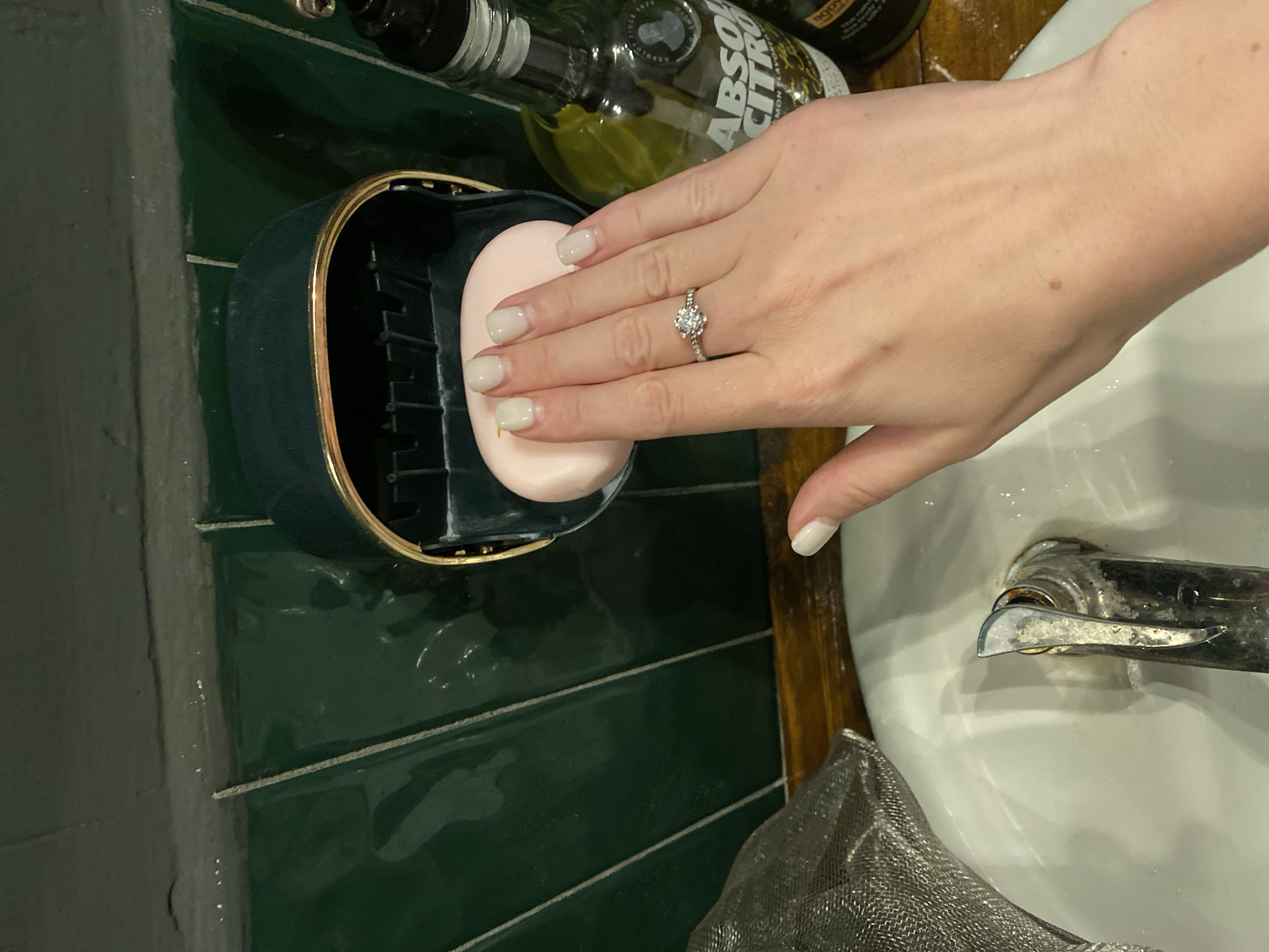 A close-up shot shows a person's hand holding a pink bar of soap in a dark green soap dish with a gold trim, positioned above a white sink. The background features green tiled walls and a wooden surface. To the right, a bottle of Absolut Citron vodka is visible. The person is wearing a diamond ring on their left ring finger and has neatly manicured nails. The scene appears to be a bathroom or kitchen sink area, but no specific location in Đà Nẵng, Vietnam, can be identified from the visual information provided.
