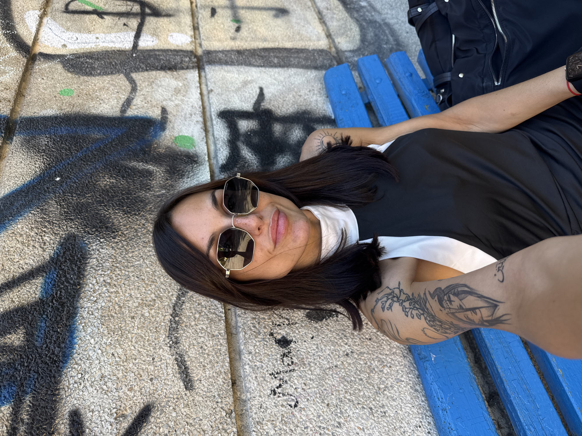 A woman is lying on a blue wooden bench in an outdoor location. The bench is situated on a concrete surface covered with graffiti. The woman is wearing sunglasses, a black top over a white collared shirt, and a black jacket. She has tattoos on her arms. The location appears to be in a city, possibly Pleven, Bulgaria, although there are no specific landmarks to confirm this. The image is taken from a slightly high angle, looking down on the scene.