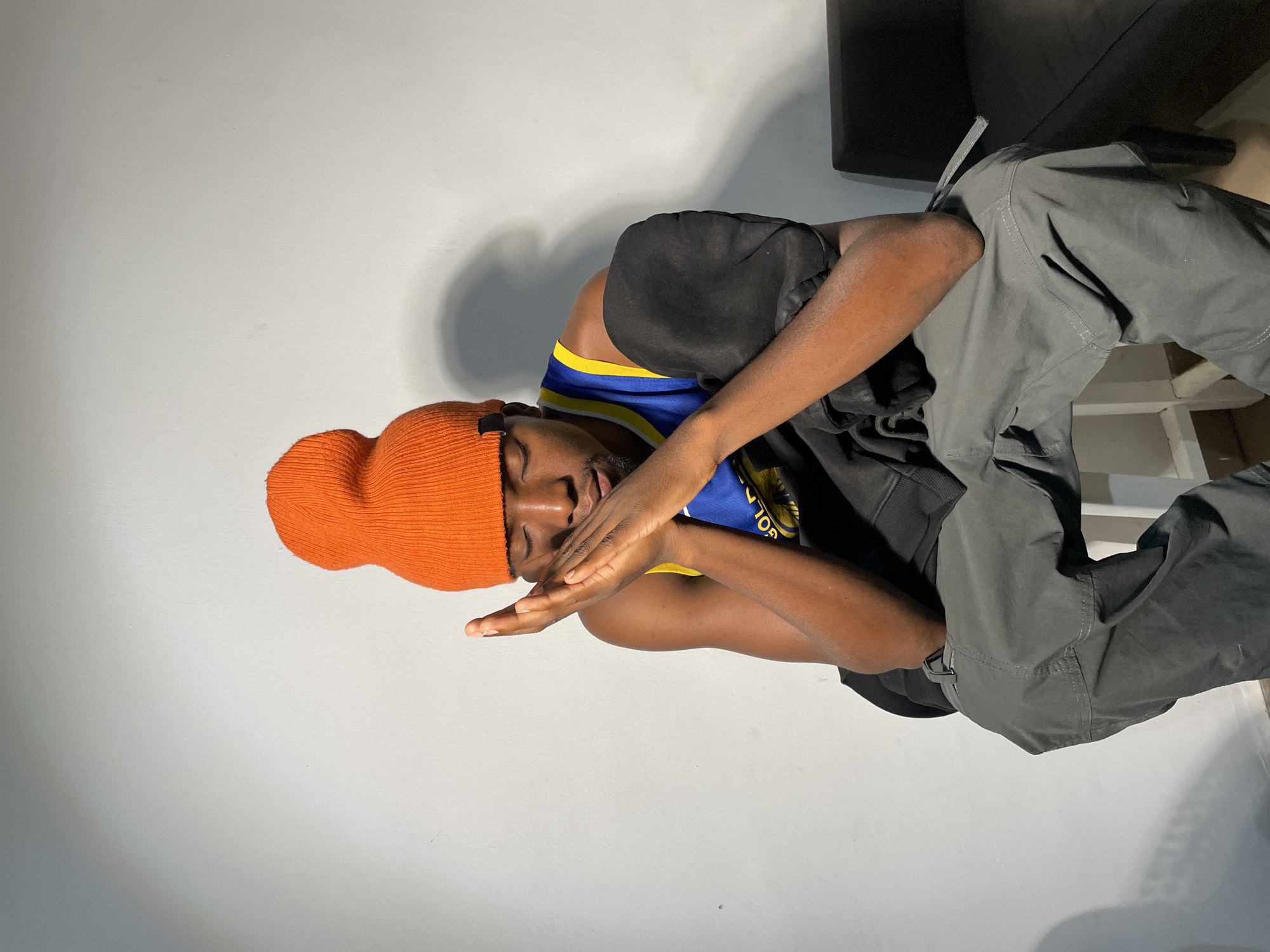 A young man with dark skin is depicted in a sideways-oriented photograph. He is wearing an orange beanie and a Golden State Warriors jersey underneath a dark-colored sweatshirt. He is also wearing gray pants. His hands are positioned near his face, which is turned slightly away from the camera. The background is a plain white wall. The scene does not appear to be in the city of Barakin Akawo, Nigeria.