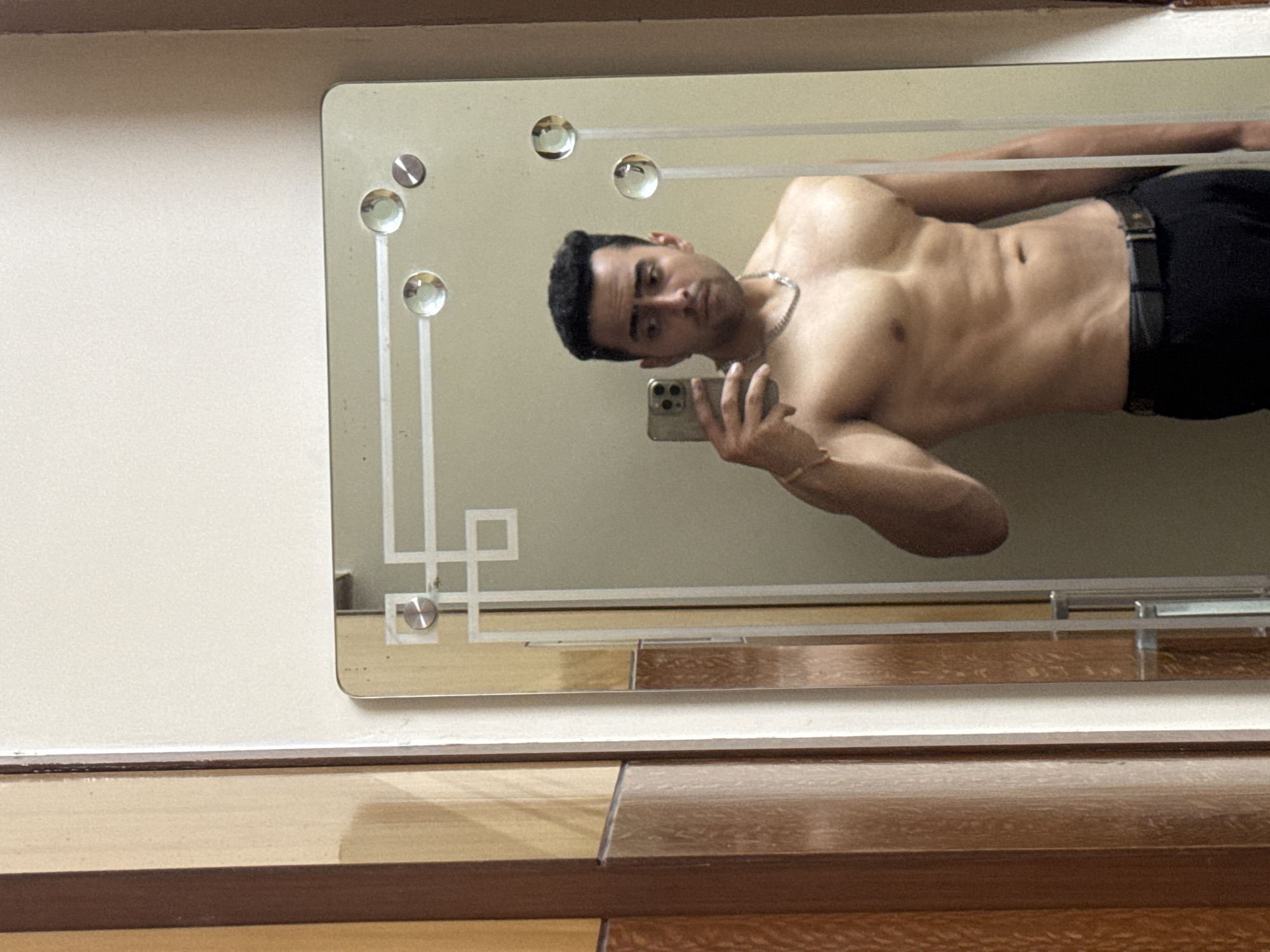 A man is seen reflected in a rectangular mirror mounted on a wall, presumably indoors. The man, shirtless and wearing black pants with a belt, holds a smartphone in his right hand, taking a selfie. He is wearing a silver chain necklace and a gold bracelet on his right wrist. The mirror is adorned with circular decorative features and geometric patterns. The wall appears to be painted in a light color. A wooden surface, possibly a table or dresser, is visible below the mirror. There is no evidence to confirm a location in Kolkata, India.