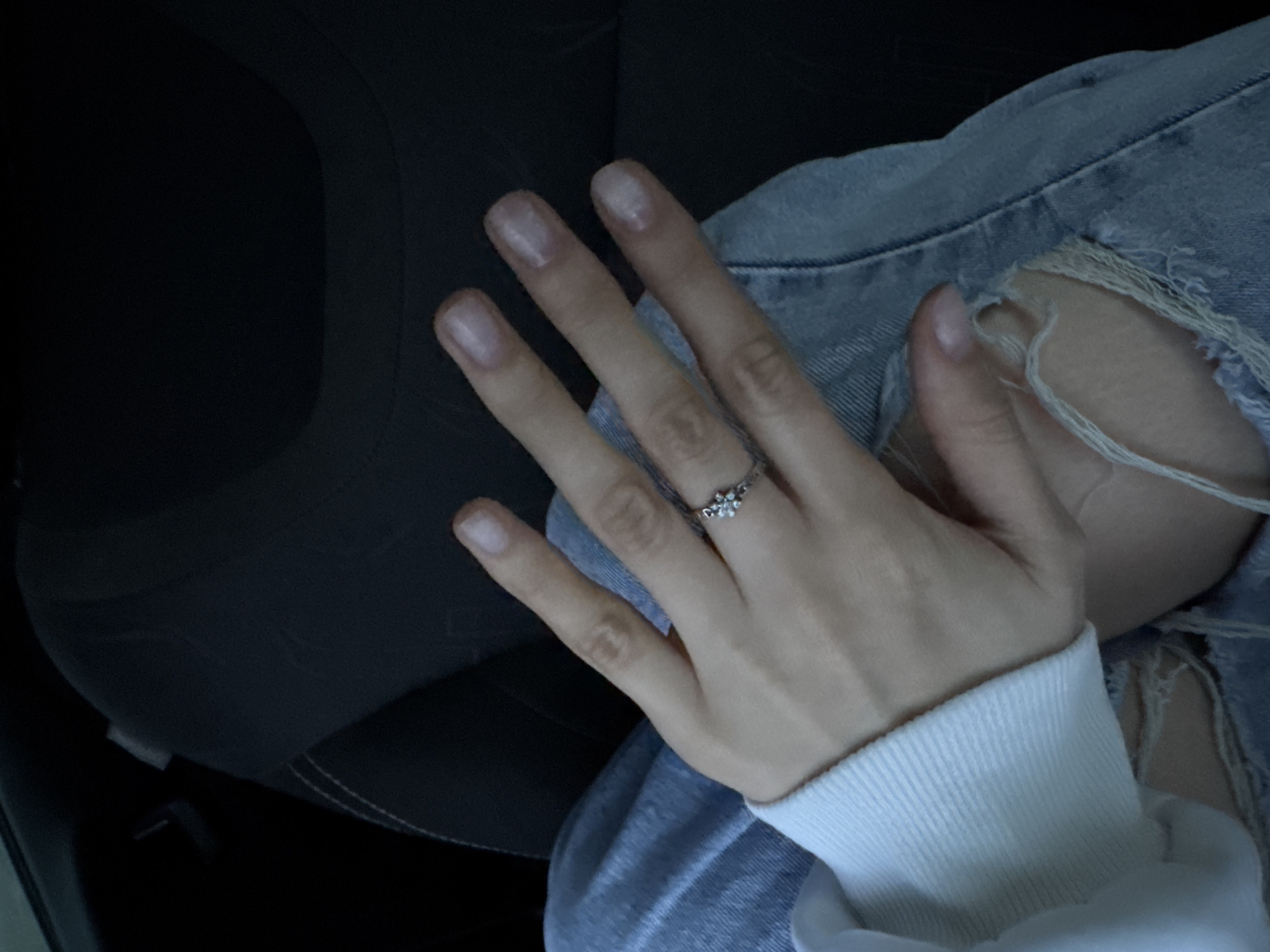 A hand, adorned with a silver ring featuring three small floral designs, rests atop a denim-clad thigh. The denim exhibits a tear, revealing frayed fabric. A white, ribbed-knit cuff encircles the wrist. The background is indistinct, revealing portions of a dark, textured surface suggesting the interior of a vehicle. The scene is not discernibly associated with Ягул, Russia.