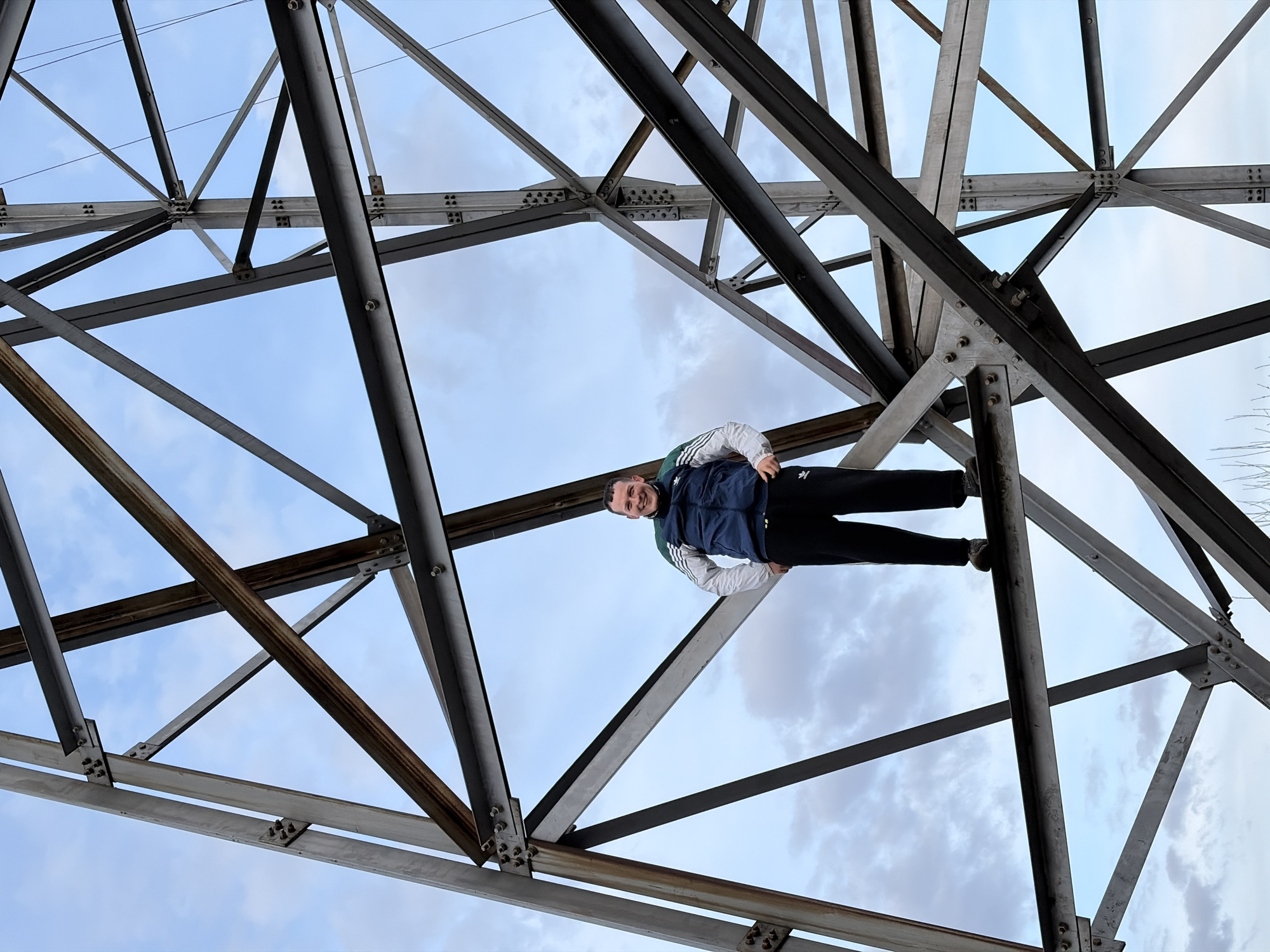 A medium shot of a man perched on the structural steel framework of a power transmission tower. He is wearing black pants and a navy blue tracksuit jacket with white stripes on the sleeves. He looks towards the camera and has a smile. The sky is pale blue with wispy clouds. This scene likely takes place in Chaykovsky, Russia, based on the prompt. The transmission tower consists of a complex arrangement of intersecting steel beams connected by bolts.