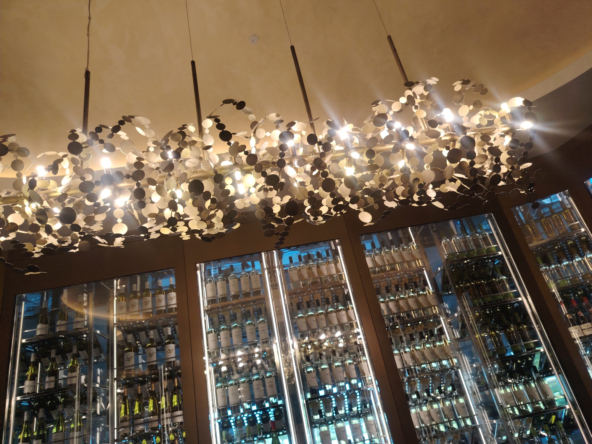 In the city of Konigil, Uzbekistan, this image captures a luxurious wine display within a modern establishment. The foreground features a multi-tiered, illuminated wine rack filled with numerous bottles of wine, predominantly white and some green. The bottles are neatly arranged on shelves with vertical LED lighting enhancing their display. Above the wine rack, a contemporary chandelier composed of numerous circular, metallic elements hangs from the ceiling, reflecting the ambient light and contributing to the sophisticated atmosphere. The ceiling is a pale, smooth surface. The background shows more shelves of wine, indicating an extensive collection. No people are visible in the scene.
