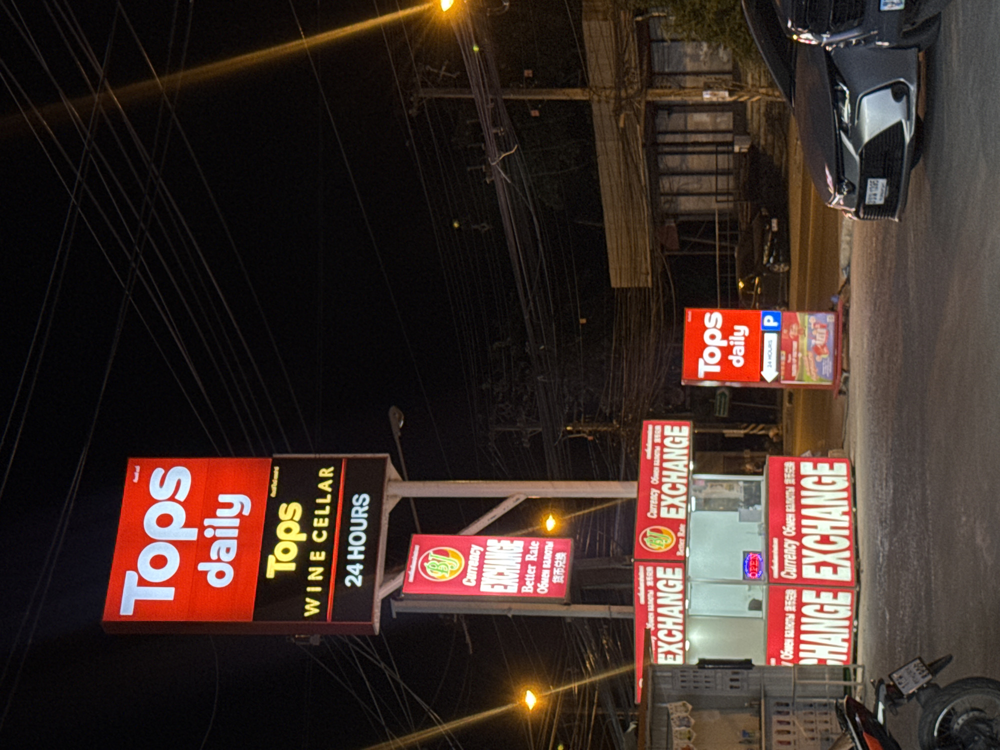 The image captures a nighttime street scene, likely in an urban setting given the presence of multiple signs and streetlights. The primary focus is on a series of illuminated signs advertising different businesses.
In the foreground, a prominent red sign displays "Tops daily" in large white lettering. To its right, another sign reads "Tops WINE CELLAR 24 HOURS." Further down the street, a smaller red sign with a yellow logo says "Currency EXCHANGE" with additional text in what appears to be a foreign script, alongside "Better Rate." This currency exchange booth is also visible, with an "OPEN" sign lit up. Adjacent to this, another large red sign repeats "EXCHANGE" in white lettering. In the background, a dark-colored car is parked, and the scene is crisscrossed with numerous overhead wires, suggesting a typical street infrastructure. The lighting from the signs casts a glow on the surroundings, and some streetlights are also visible. The overall impression is that of a commercial area operating at night. No people are clearly visible in the image. The weather is not discernible.