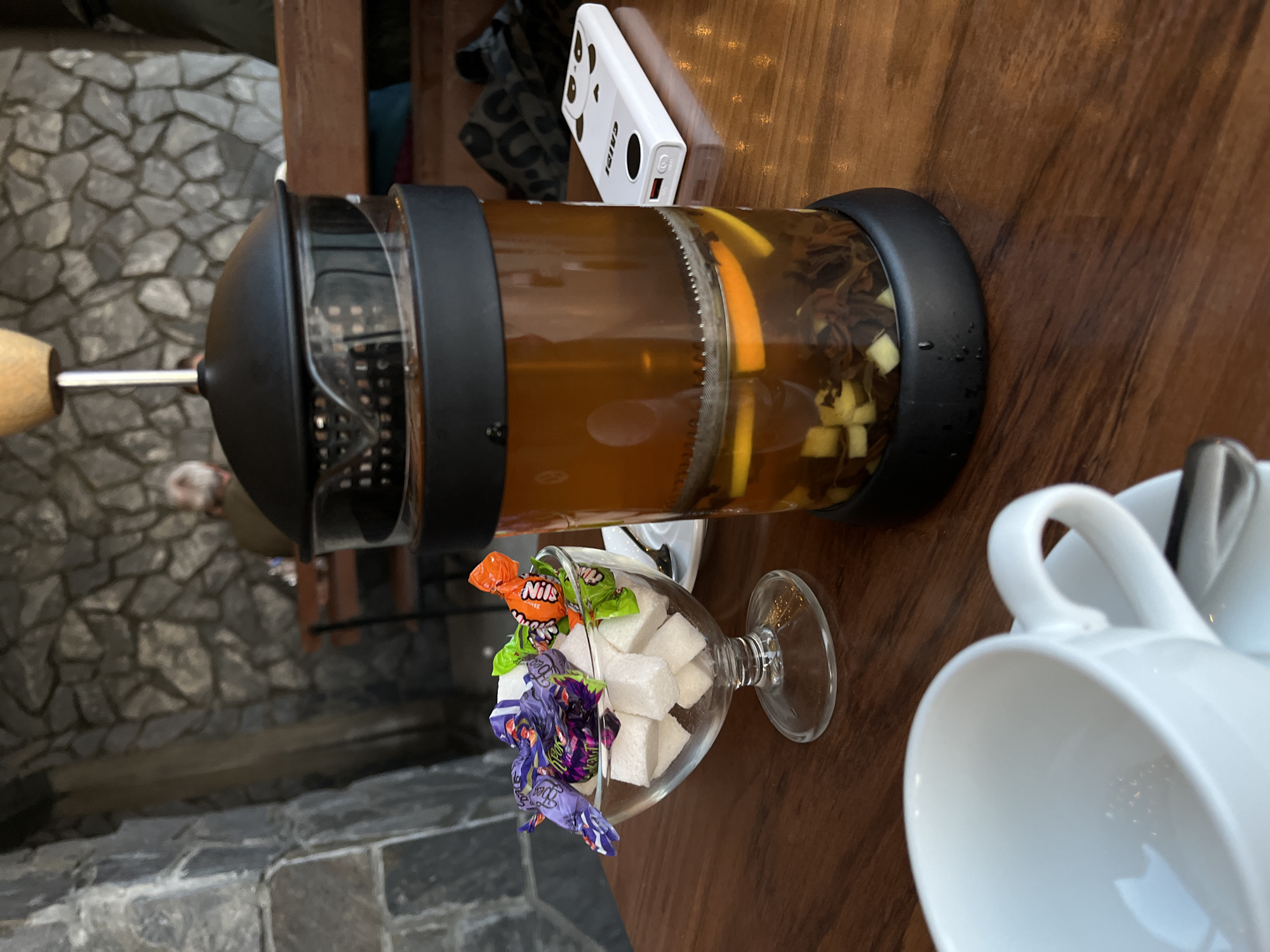 A close-up, eye-level shot captures a table setting at what appears to be a cafe in Yelizovo, Russia. The background is dominated by a rough, gray stone wall, suggesting an rustic or naturalistic interior design. A blurred figure of a person is visible in the distance, seated at another table.
In the foreground, the table is laden with items for a tea break. To the left, a clear glass coupe dish is filled with white sugar cubes and topped with several individually wrapped candies, their colorful wrappers hinting at fruity or sweet fillings. The candies are purple, green, and orange.
Dominating the center of the frame is a glass French press containing brewed tea. The tea is a deep amber color and contains loose tea leaves, slices of orange, and small, diced pieces of what might be apple or pear. The French press has a black lid and base, with a metal plunger assembly visible within the glass cylinder.
To the right of the French press, a white smartphone with a black panda face design is placed on the wooden table. The phone is lying flat, displaying a charging port.
In the lower left corner, a white ceramic teacup and saucer are partially visible, suggesting that the tea is ready to be poured. The overall impression is one of a relaxed break, with elements of both indulgence (sweets) and refreshment (tea). The lighting is moderate, with some highlights reflecting off the glass and ceramic surfaces.