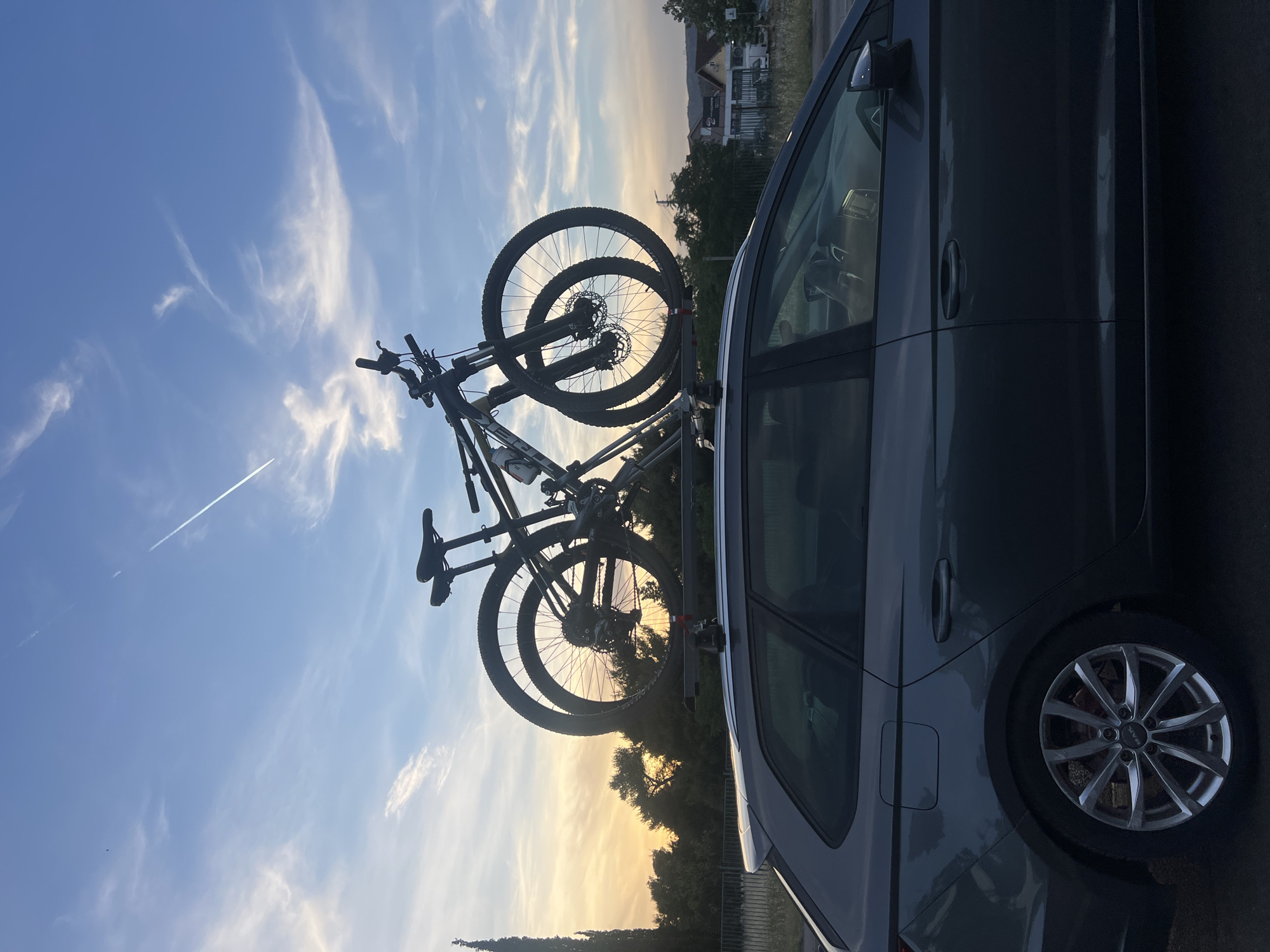 The image shows a gray car parked with two bicycles mounted on a roof rack. The background features a sky with wispy clouds and the setting sun casting a warm glow. Trees and a building are faintly visible in the distance. The location appears to be outdoors, possibly in a residential area, based on the visible buildings and trees, though the specific location within Podgorica, Montenegro, is unclear without additional visual cues or contextual information. The main subjects are the car and the bicycles, with the car acting as the primary transport for the two bicycles which are both mountain bikes.