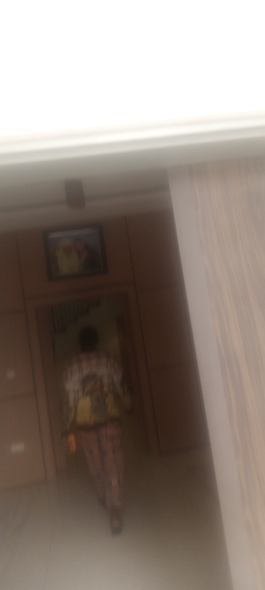 A blurry, low-quality image shows a person standing in what appears to be a hallway or entryway. The person is wearing a patterned shirt and pants. Behind the person is a wood-paneled wall with a framed picture hanging above. To the right of the scene is a blurred vertical stripe. The location is presumed to be in Unguwar Rimi, Nigeria.