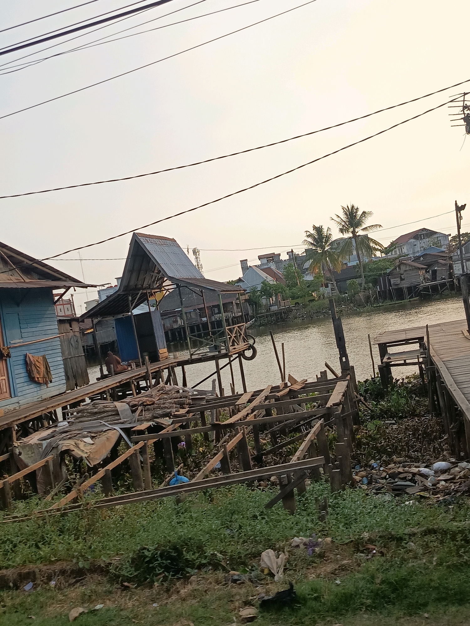 The image depicts a waterfront scene in Banjarmasin, Indonesia, characterized by stilt houses and a wooden walkway extending over a body of water. The houses are a mix of weathered wood and some appear to have corrugated metal roofs. One house on the left is painted light blue. In the foreground, there is an unfinished wooden structure with lumber stacked on it, and debris including plastic bottles and wrappers is visible on the grassy bank. A person is seated on a wooden platform in front of one of the stilt houses. Palm trees are visible in the background, along with other residential structures further down the river. The sky is overcast, suggesting either early morning or late afternoon light. The overall impression is one of a lived-in, somewhat cluttered, but functional riverside community.