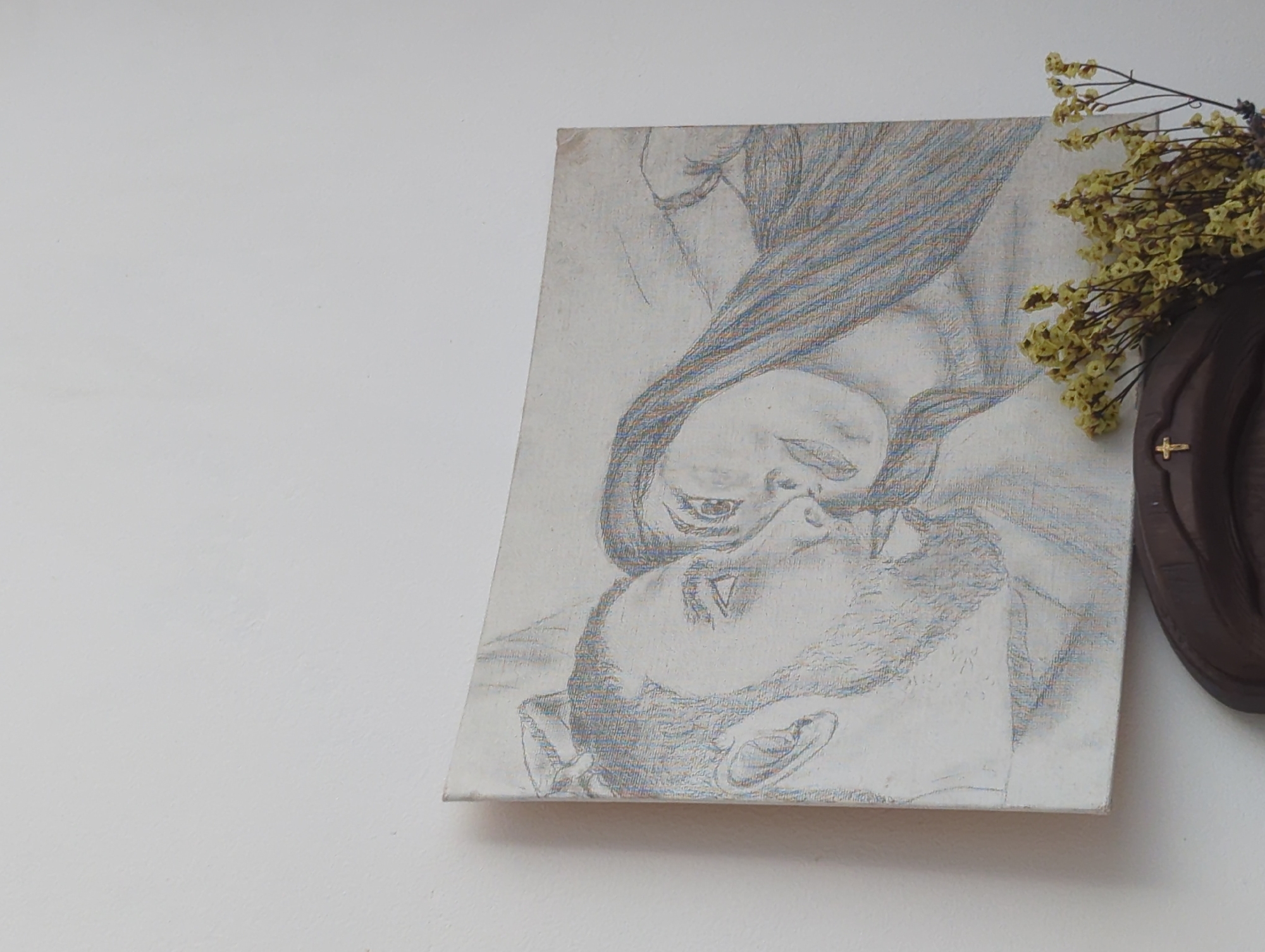 The image depicts a drawing on a canvas board, positioned against a plain, light-colored wall in Burgas, Bulgaria. The drawing, executed with colored pencils, illustrates a man and a woman in close proximity, facing each other. The man, on the left, is rendered in profile, featuring a beard and short hair, looking towards the woman. The woman, on the right, is shown in a three-quarter profile with long, flowing hair delineated with blue and reddish lines, her gaze directed towards the man. Visible pencil strokes and canvas texture are evident throughout the artwork.
Below the drawing, a portion of a dark brown, possibly wooden, decorative object is visible, featuring a small, gold-colored cross on its left side. A cluster of dried flowers, predominantly yellow-green, is arranged atop this object, partially obscuring the lower right corner of the canvas. The background is an unadorned white wall, showing minor surface irregularities.